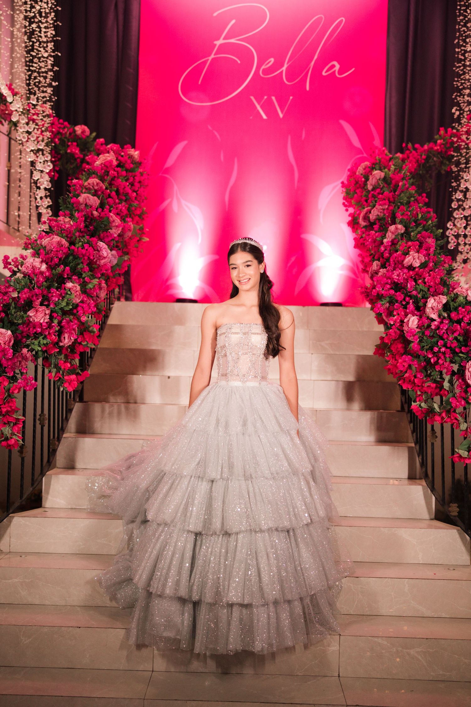 A stunning quinceañera celebration with a sparkly silver ballgown against a pink-lit backdrop with red floral arrangements.