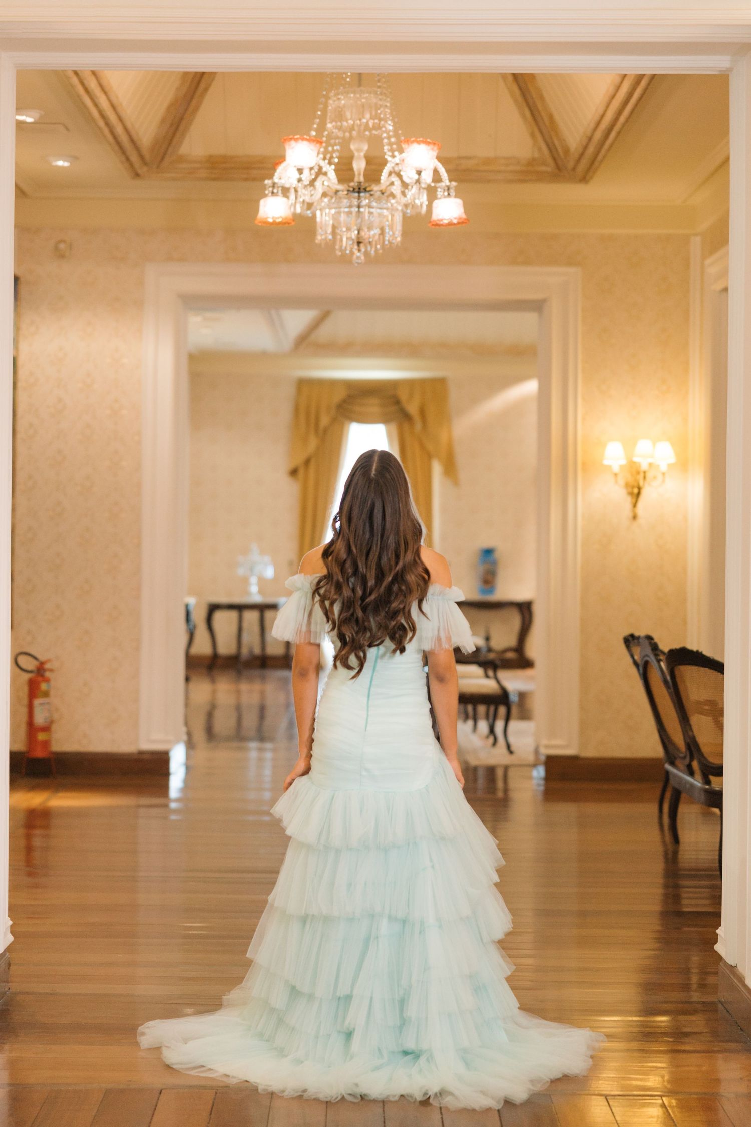A mint green ruffled gown photographed from behind in an elegant hallway with crystal chandeliers.
