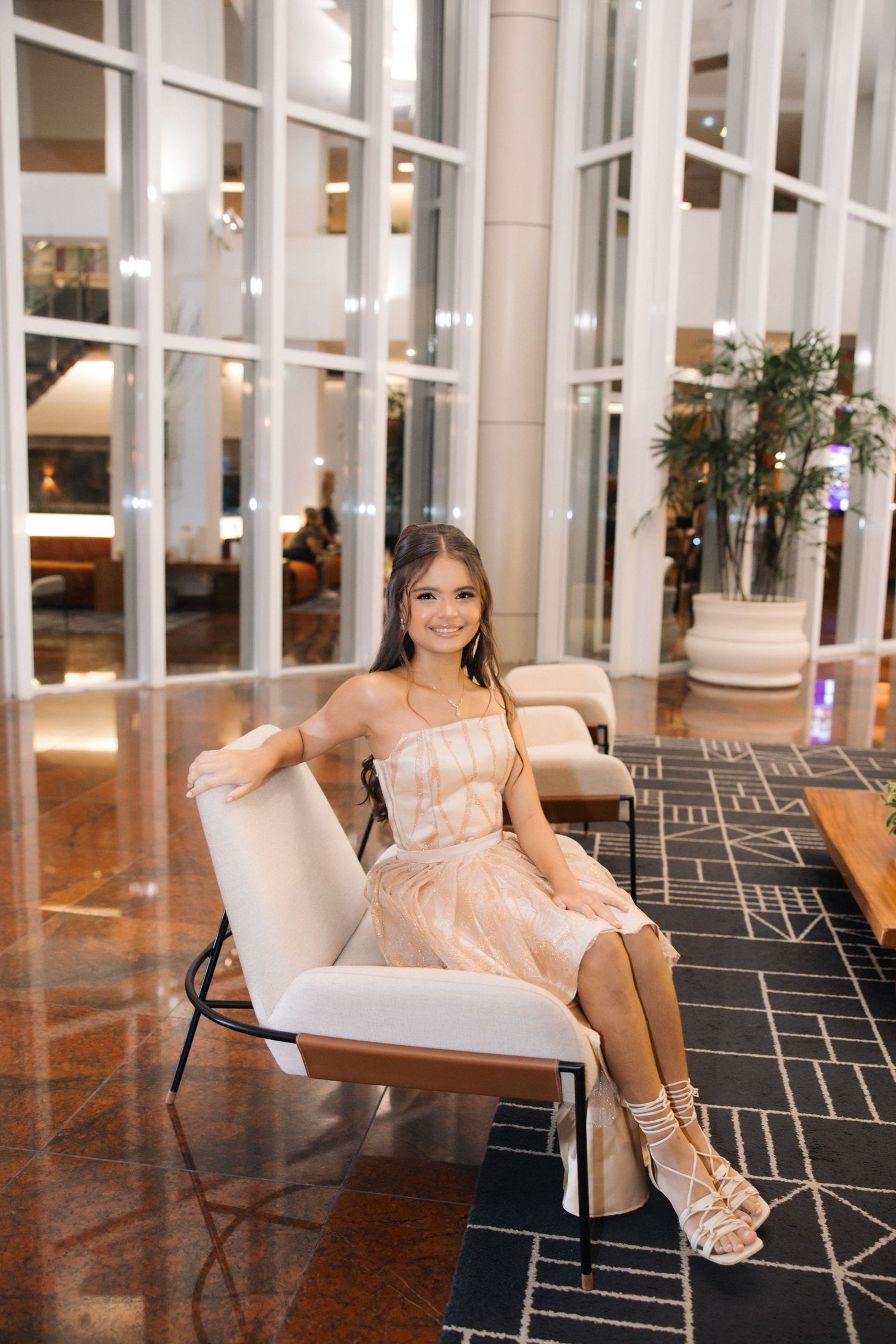 A woman in a peach-colored dress sits elegantly on a white lounge chair in a modern lobby with floor-to-ceiling windows.