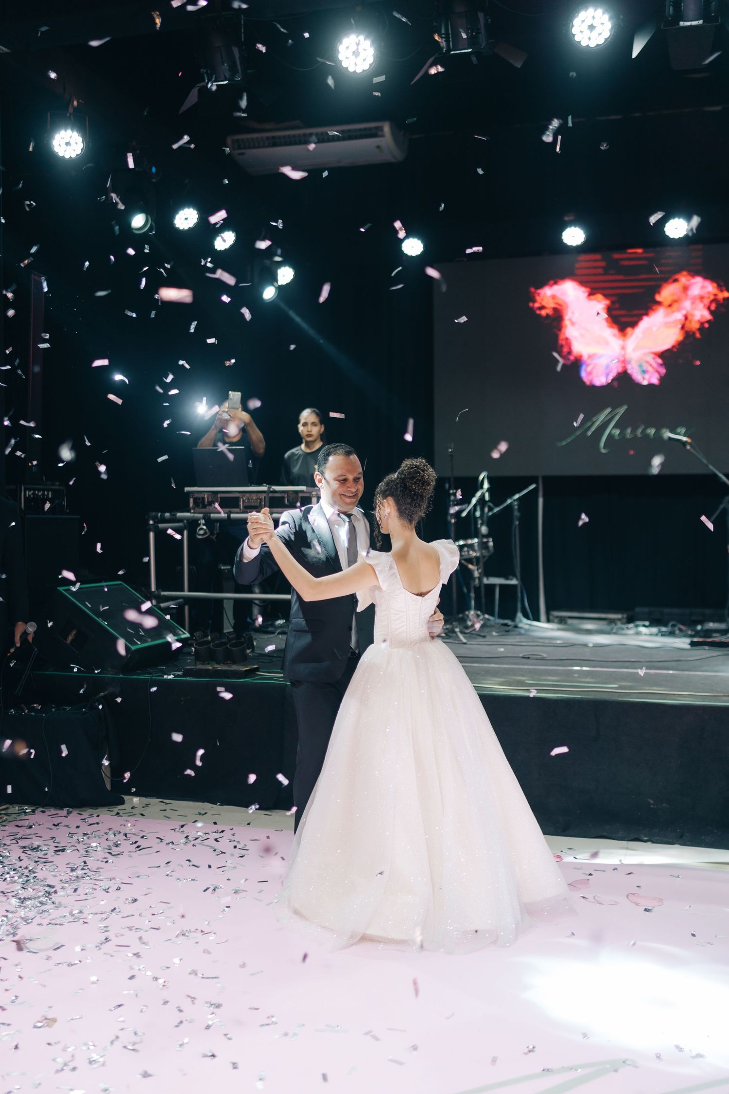 A couple shares their first dance at a wedding reception with falling confetti and a pink butterfly projection on screen.