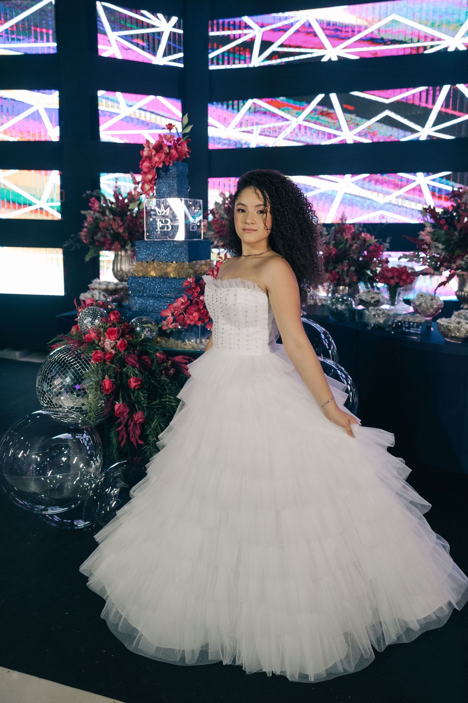 A stunning white tulle ballgown is displayed next to a cake with red floral decorations against a geometric LED backdrop.