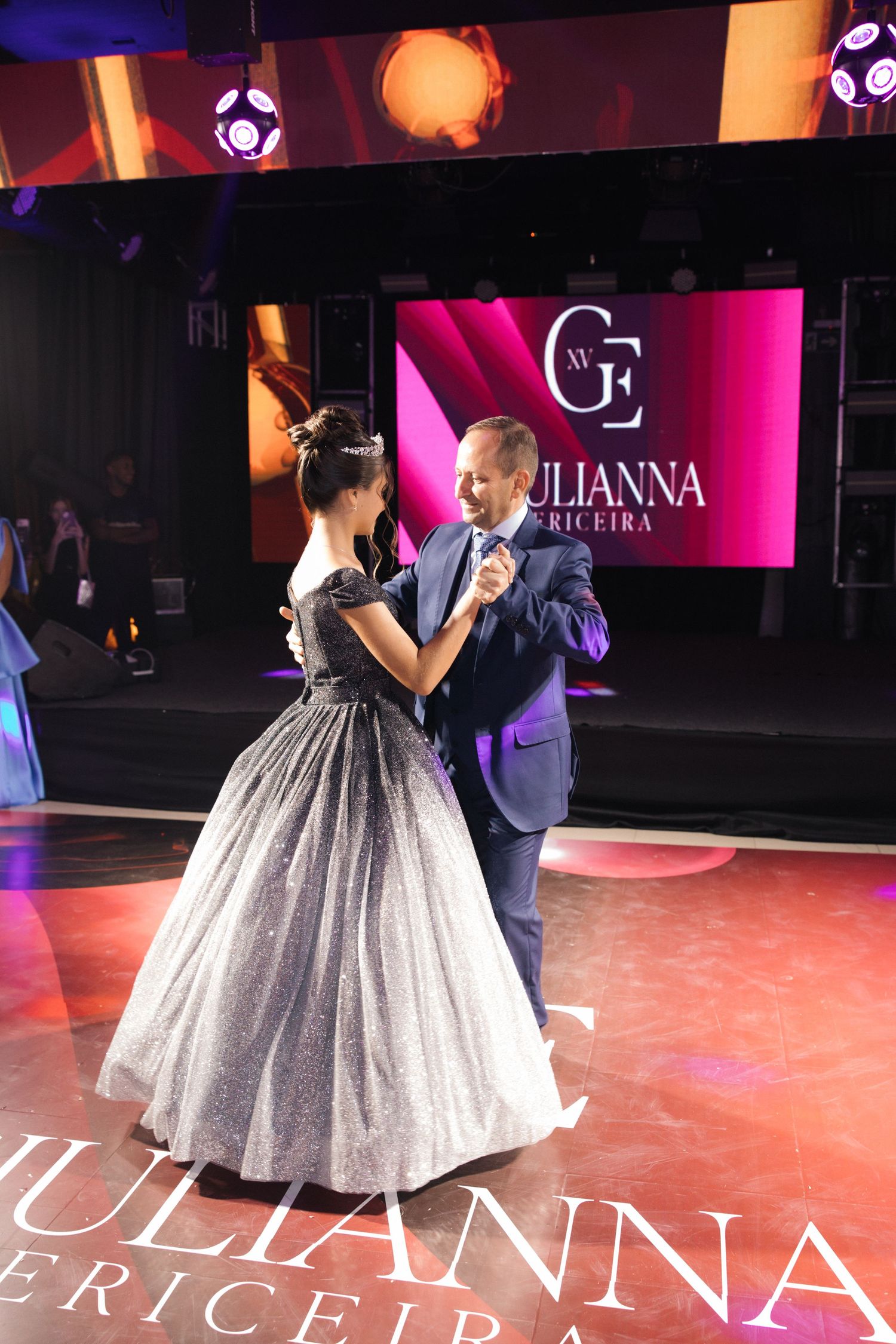 A couple dancing at an event with a sparkly ball gown against pink-lit backdrop.
