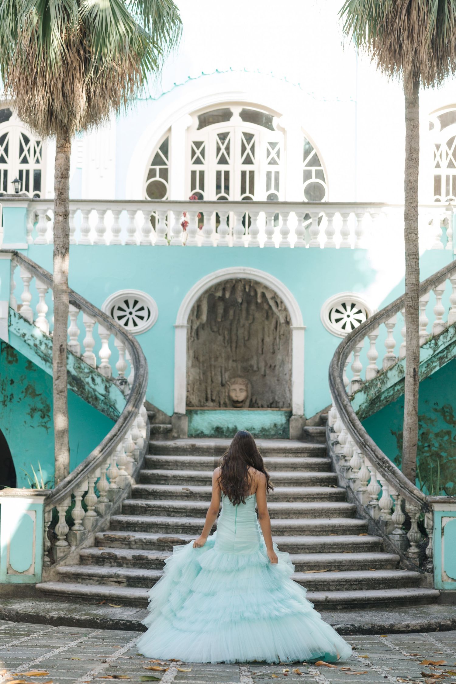 Woman in a turquoise ballgown stands at the base of an ornate staircase with palm trees and aqua architecture.