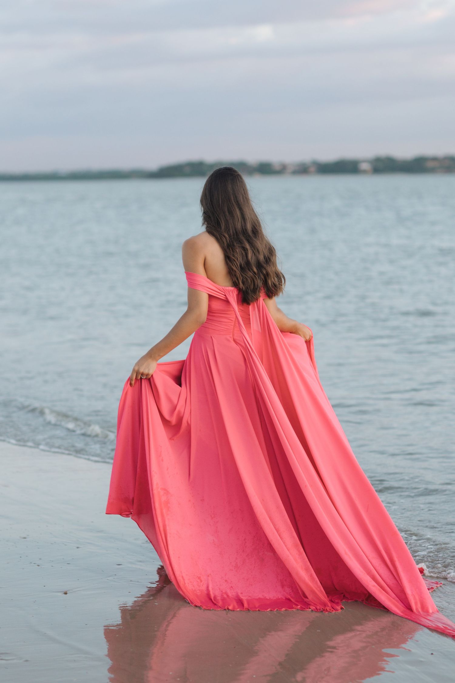 Coral flowing gown silhouetted against ocean waves at sunset on sandy beach.