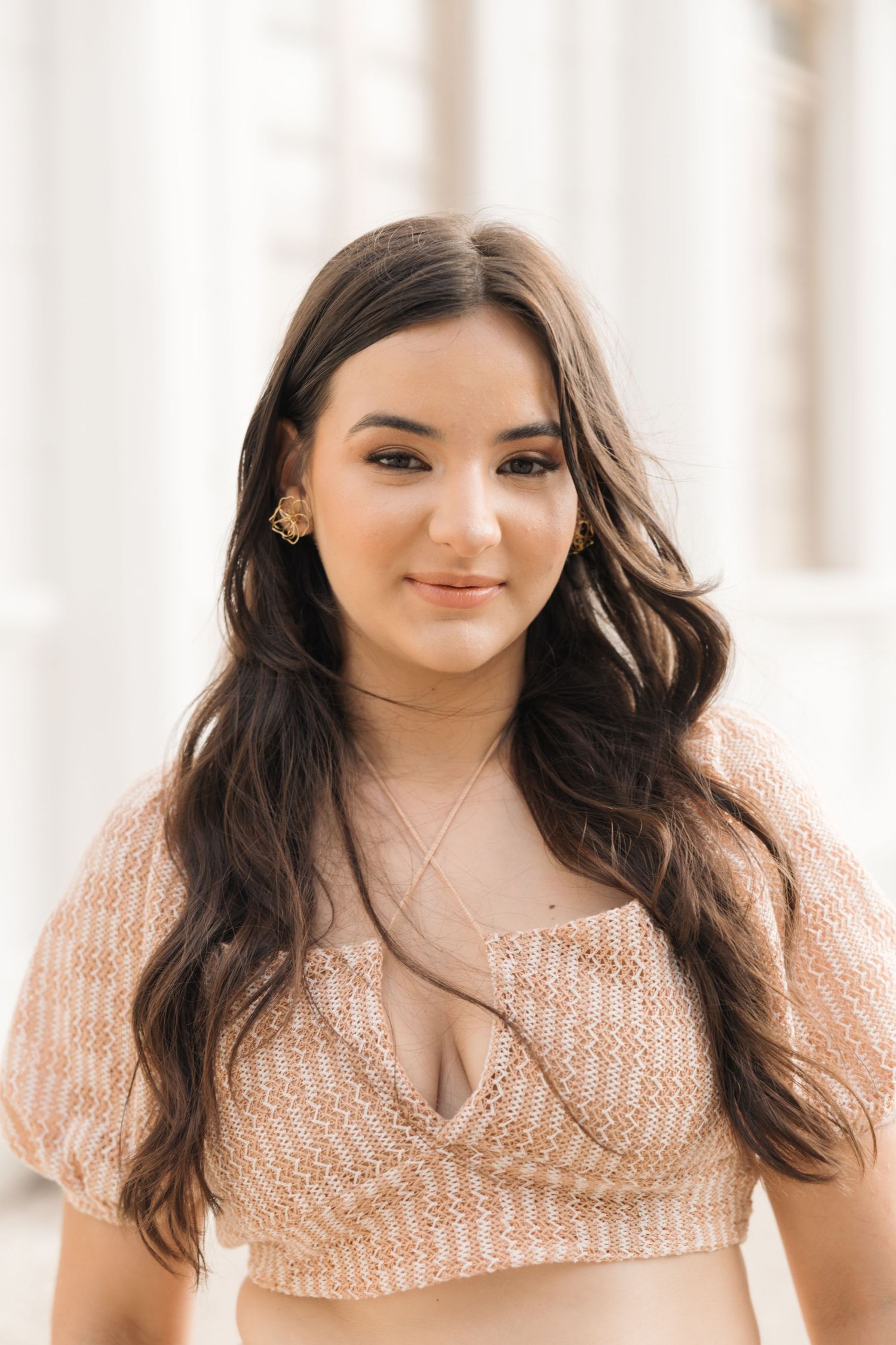 Woman in peach-colored crop top poses outdoors with long dark wavy hair and natural makeup.