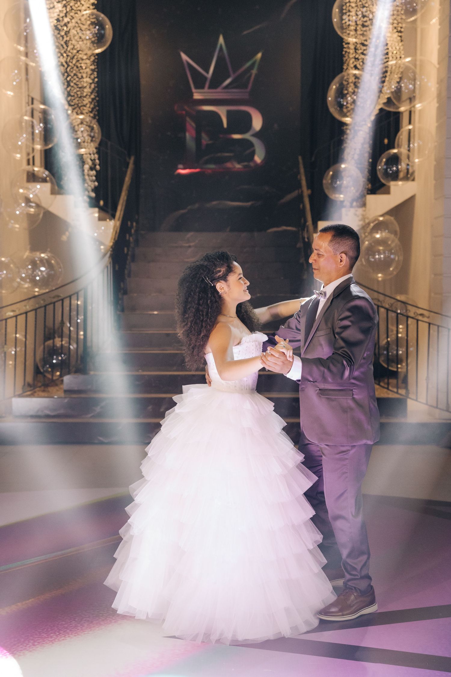 A couple dances at a formal event under decorative lighting and a crown logo on the wall.