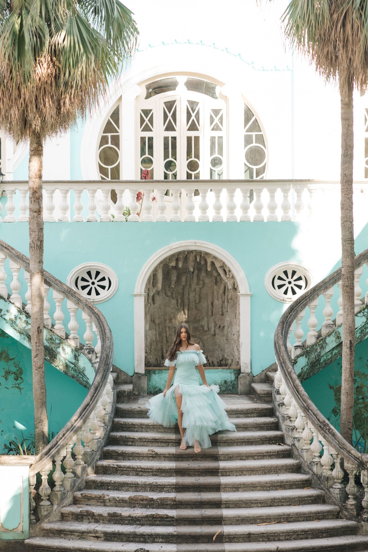 Dreamy aqua dress flows down ornate white staircase framed by palm trees and turquoise architecture.