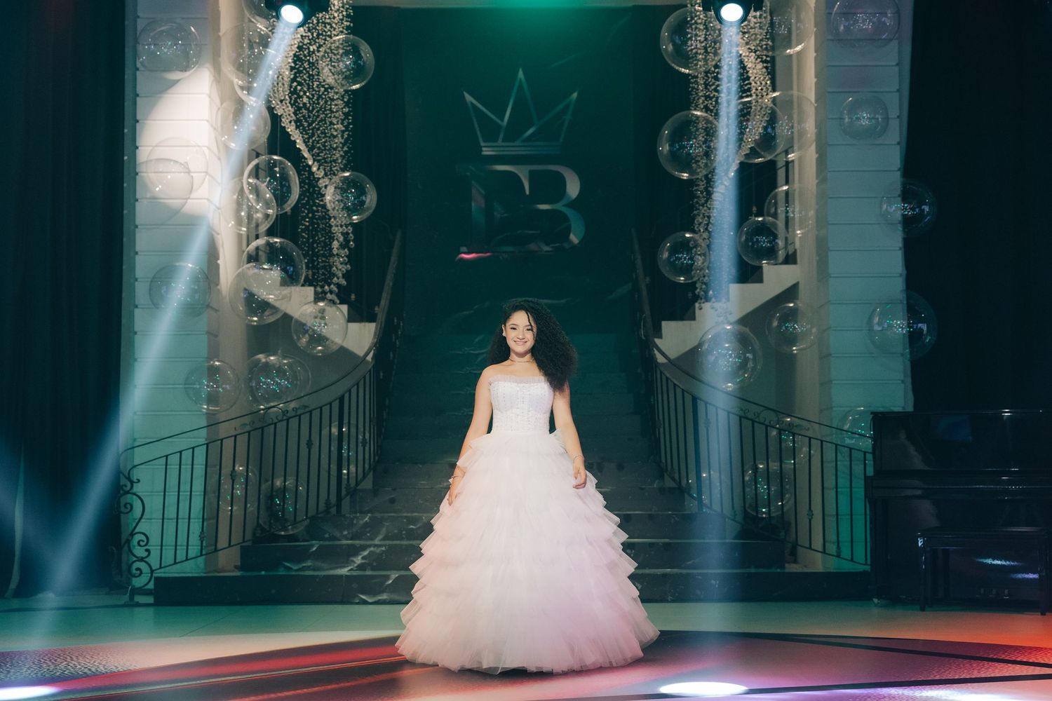 A person in a flowing white ball gown stands on an illuminated stage with ornate decorative elements and theatrical lighting.