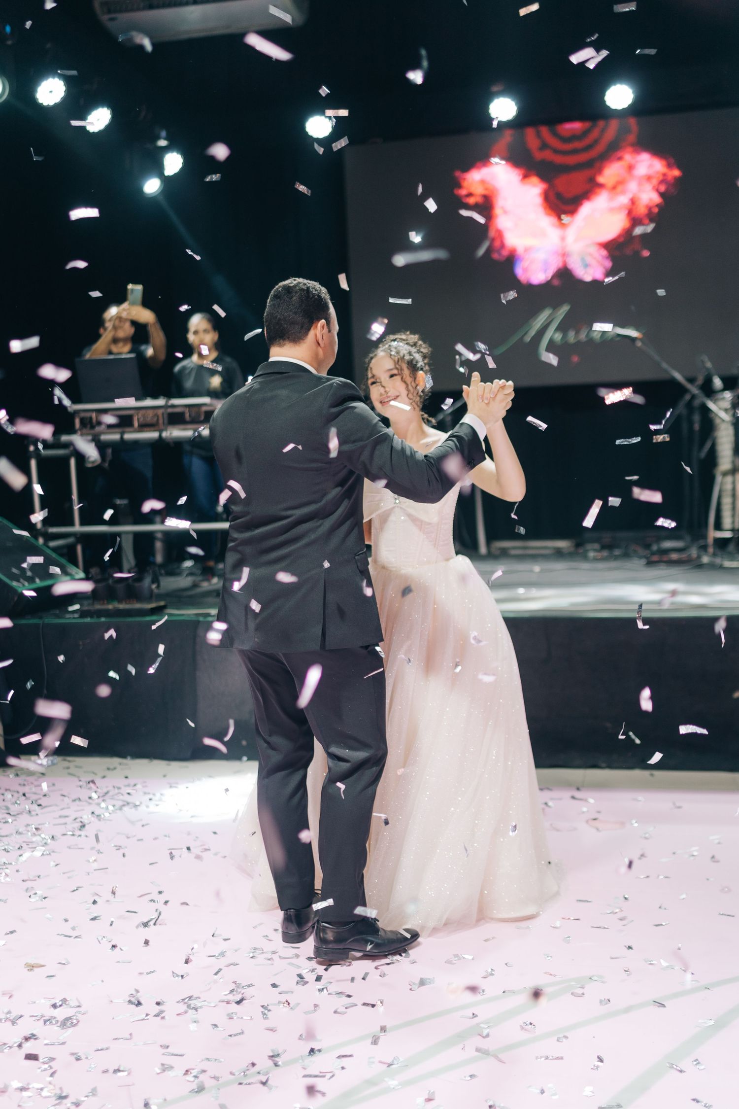 A couple dances their first dance surrounded by falling confetti under stage lighting at their wedding reception.