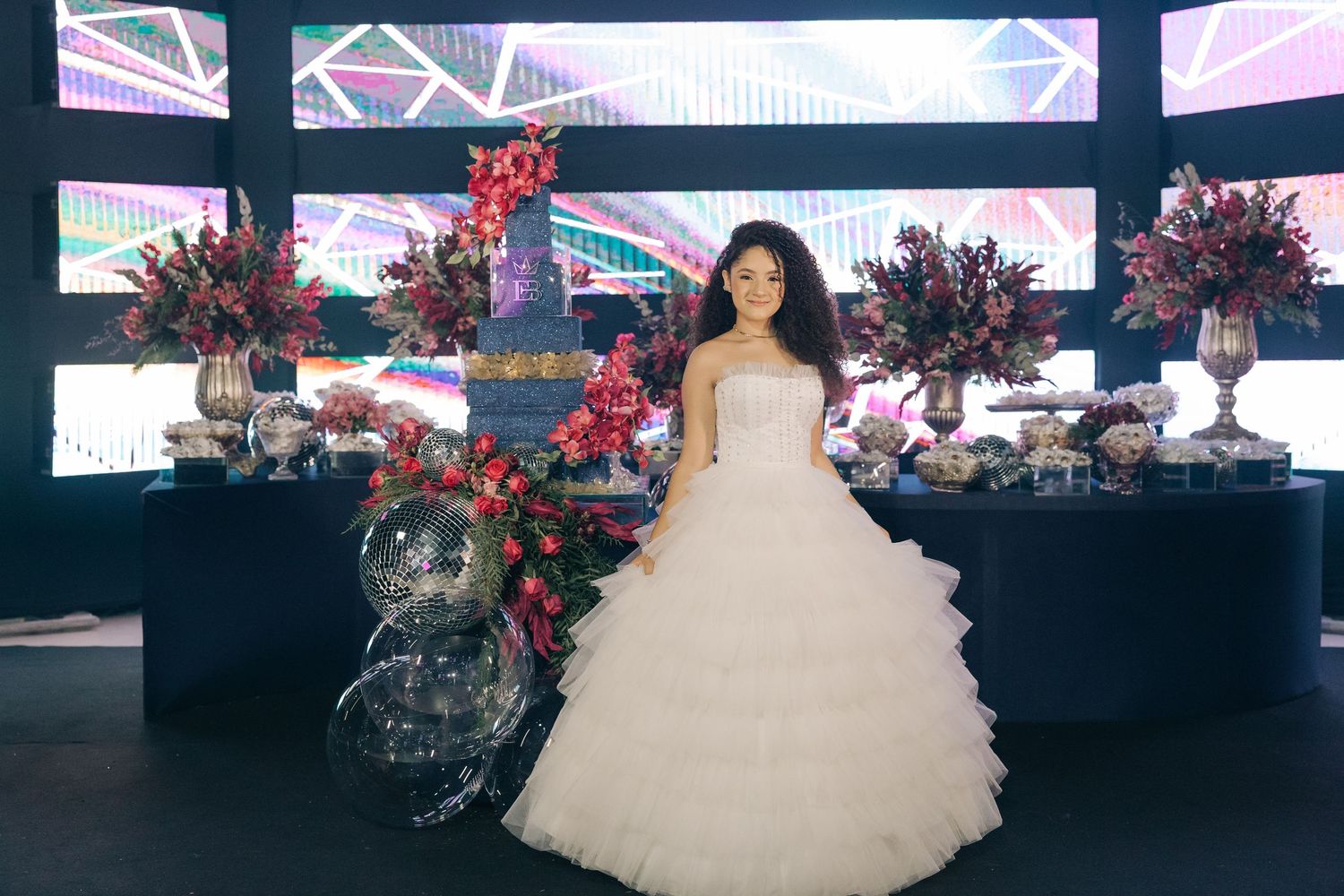 A figure in a full white ballgown stands in front of a modern LED wall display with red floral arrangements and black decor.