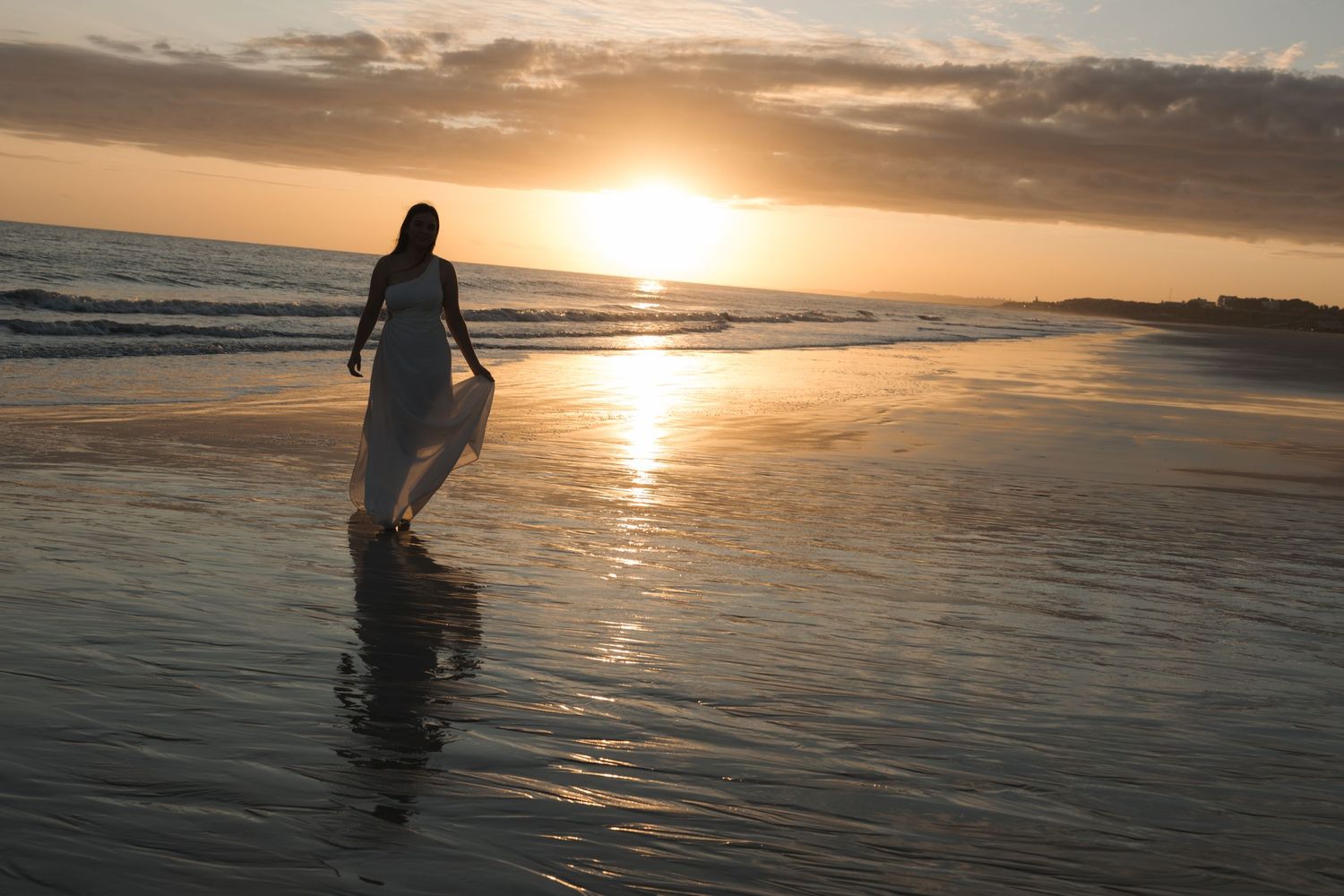 A silhouette walks along a reflective beach at sunset with golden light shimmering on the wet sand and ocean waves.