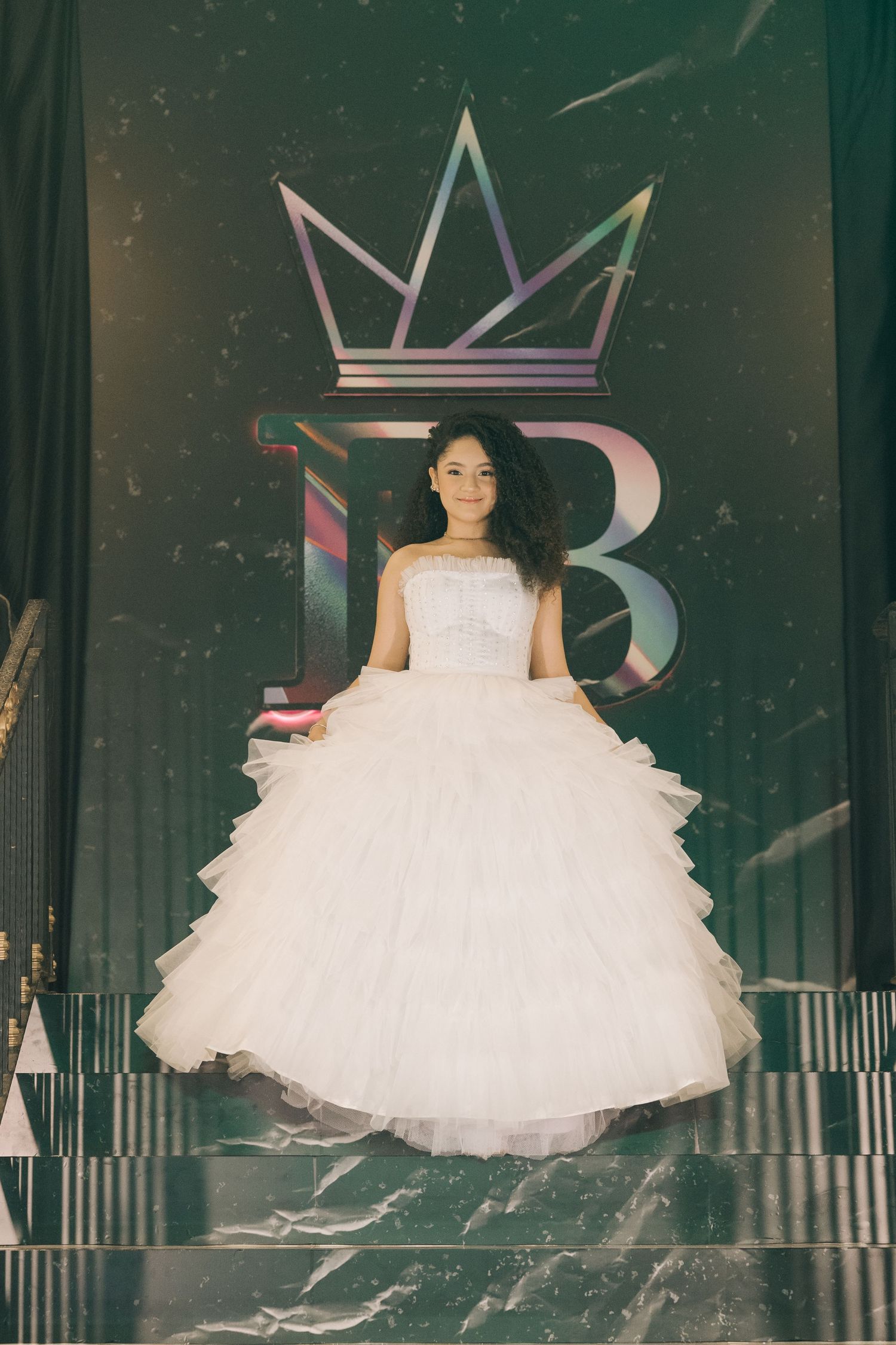 A white ruffled ballgown photographed against a dramatic backdrop with crown logo and lighting effects.