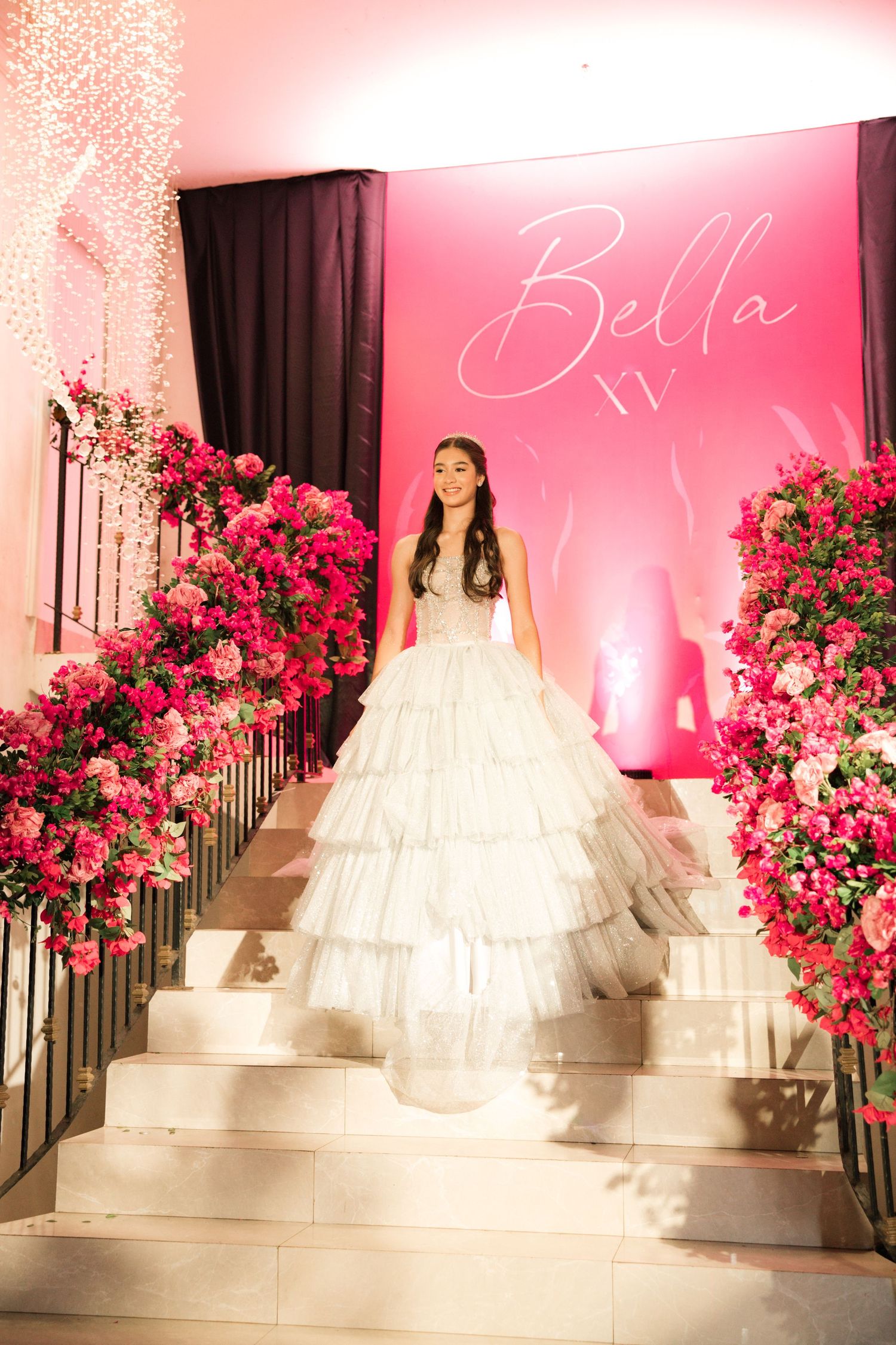 A figure in a tiered white ballgown stands on marble stairs surrounded by pink floral arrangements and a neon Bella sign.