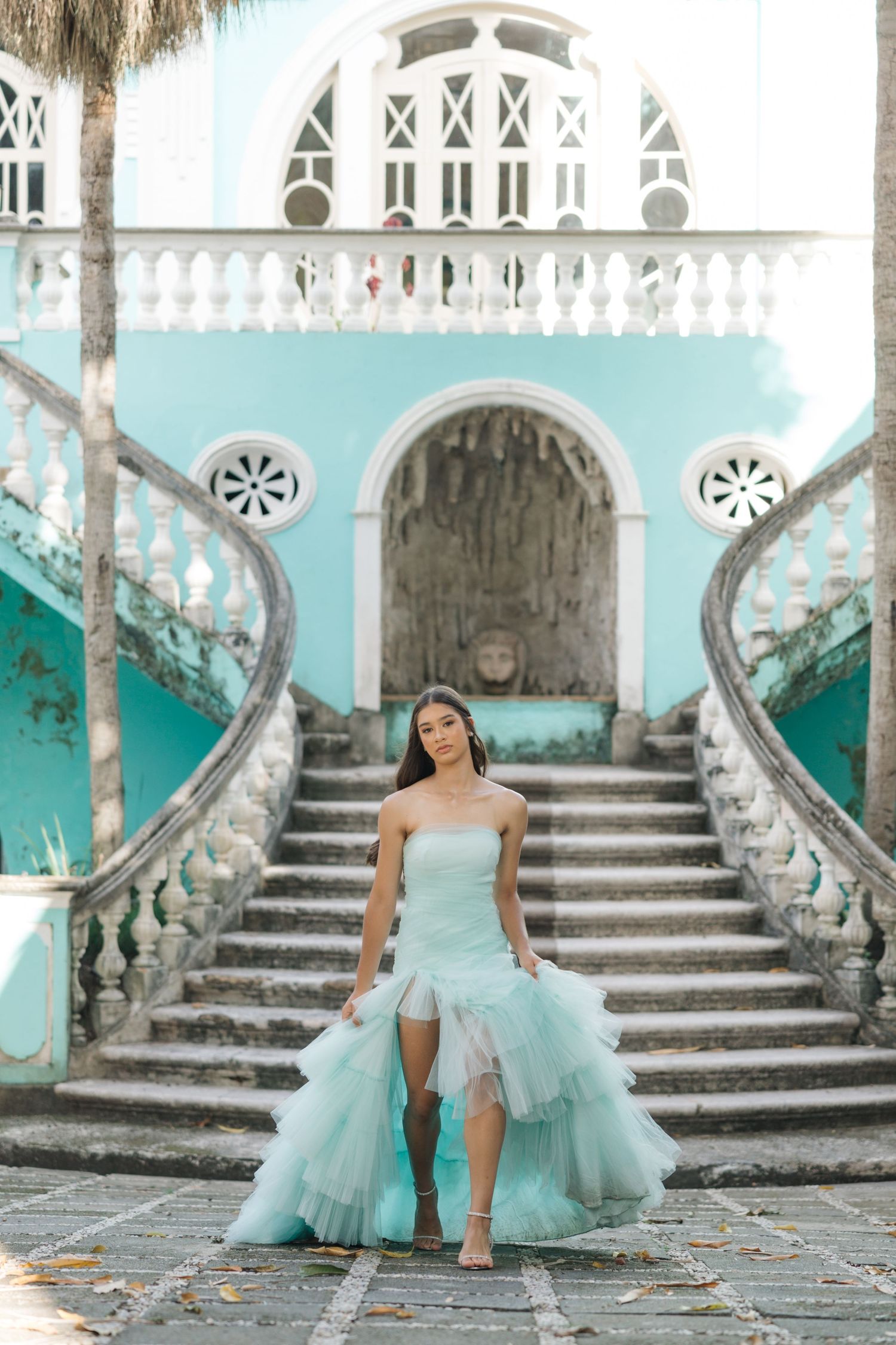 An ethereal light blue gown with cascading ruffles is featured on grand turquoise stairs with ornate architecture.