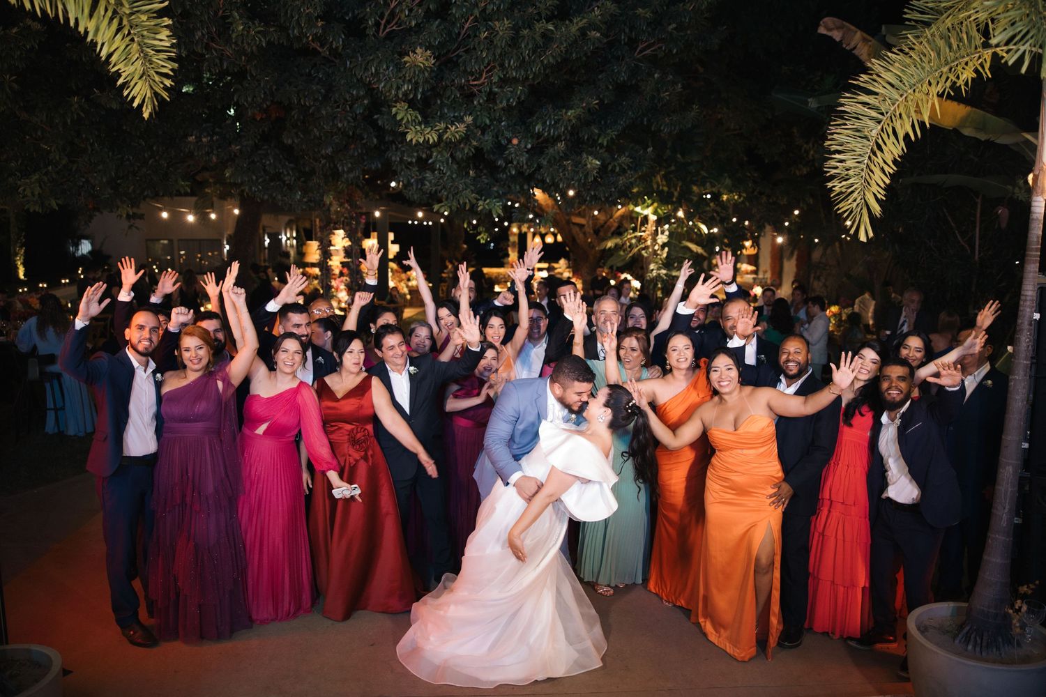 Large wedding party dressed in colorful formal attire poses with upraised arms during evening celebration.