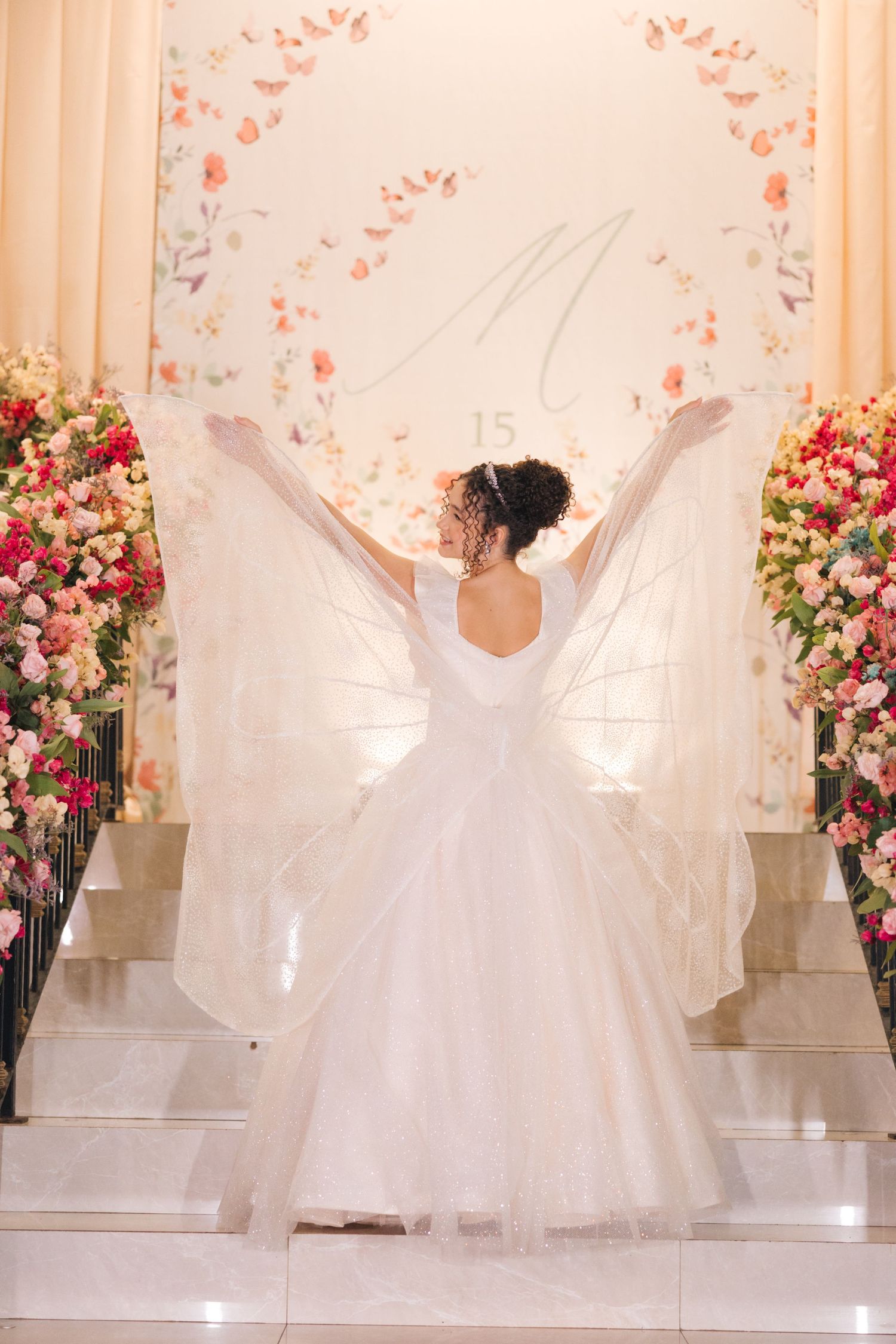 A bride in a white ballgown twirls on a dance floor surrounded by pink floral arrangements and light curtains.