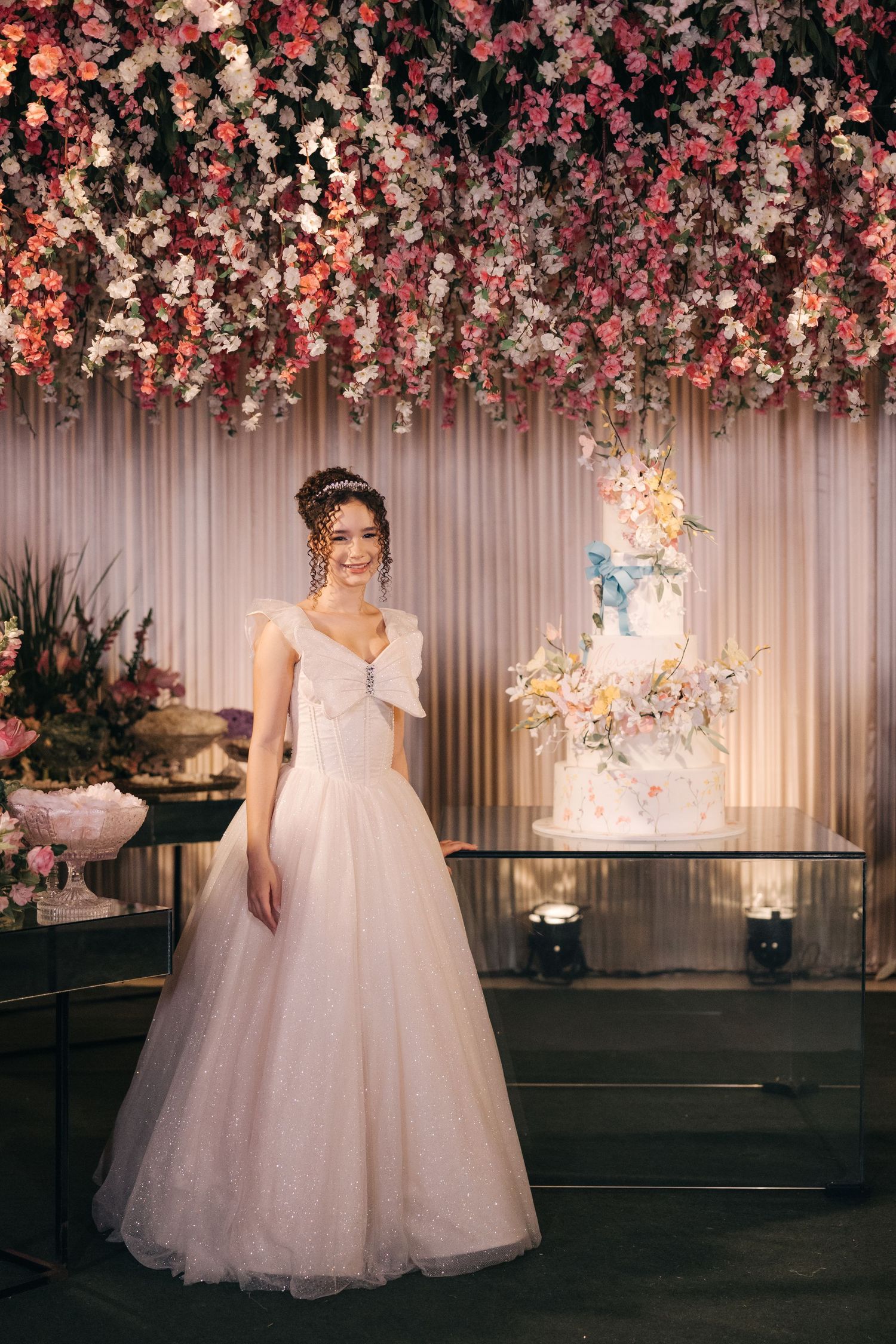 An elegant wedding reception setup with pink floral ceiling decor and a multi-tiered illuminated cake display.