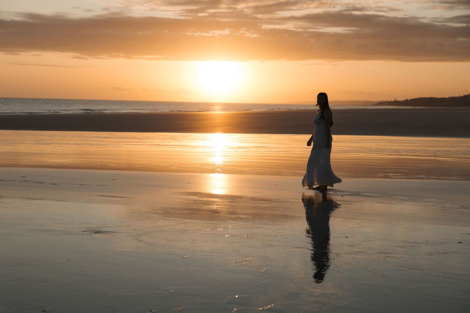 Silhouette of person walking on reflective beach at sunset with golden light dancing across wet sand.