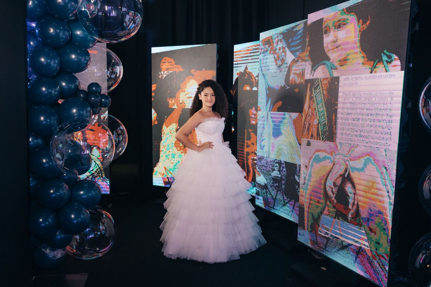 A figure in a lavish white tulle ballgown stands beside illuminated display screens with blue balloon decorations.