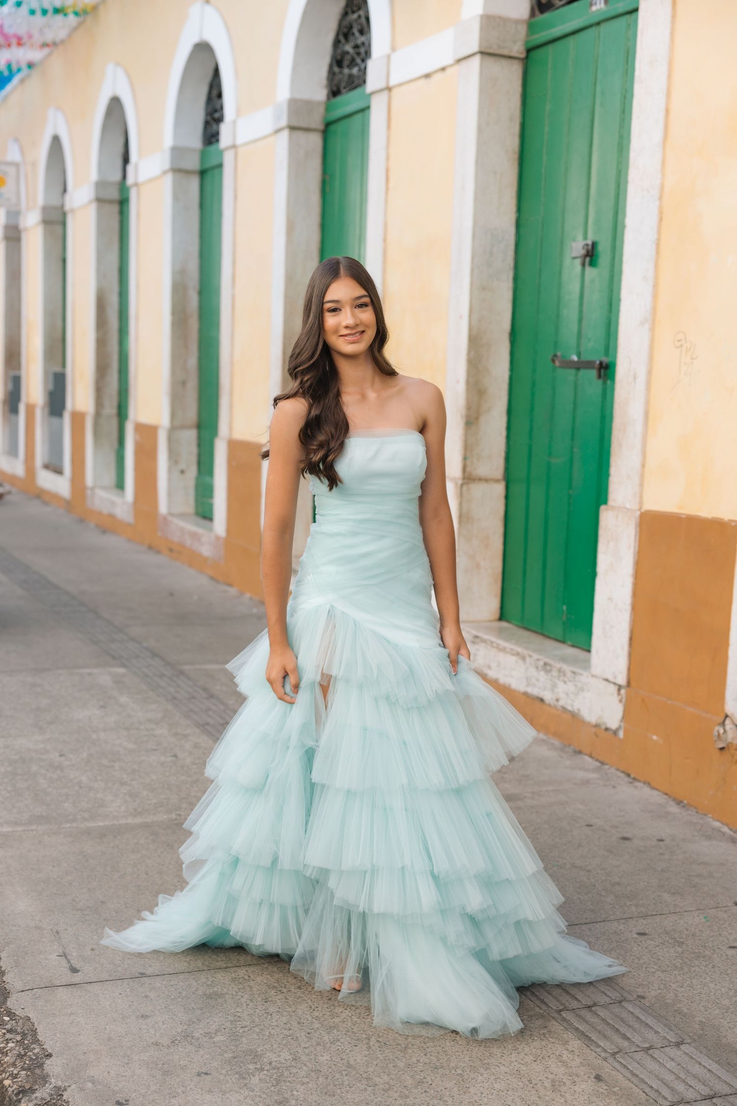A strapless light blue tulle gown with tiered ruffles stands out against a colonial building with green doors and arches.