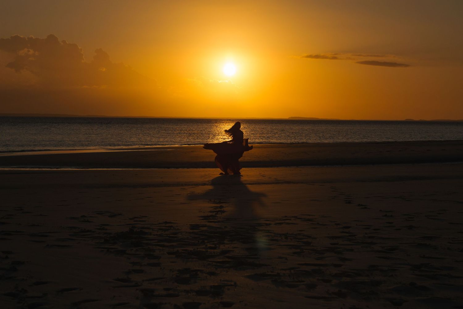 A silhouetted figure poses on a beach during a golden sunset over the ocean horizon.