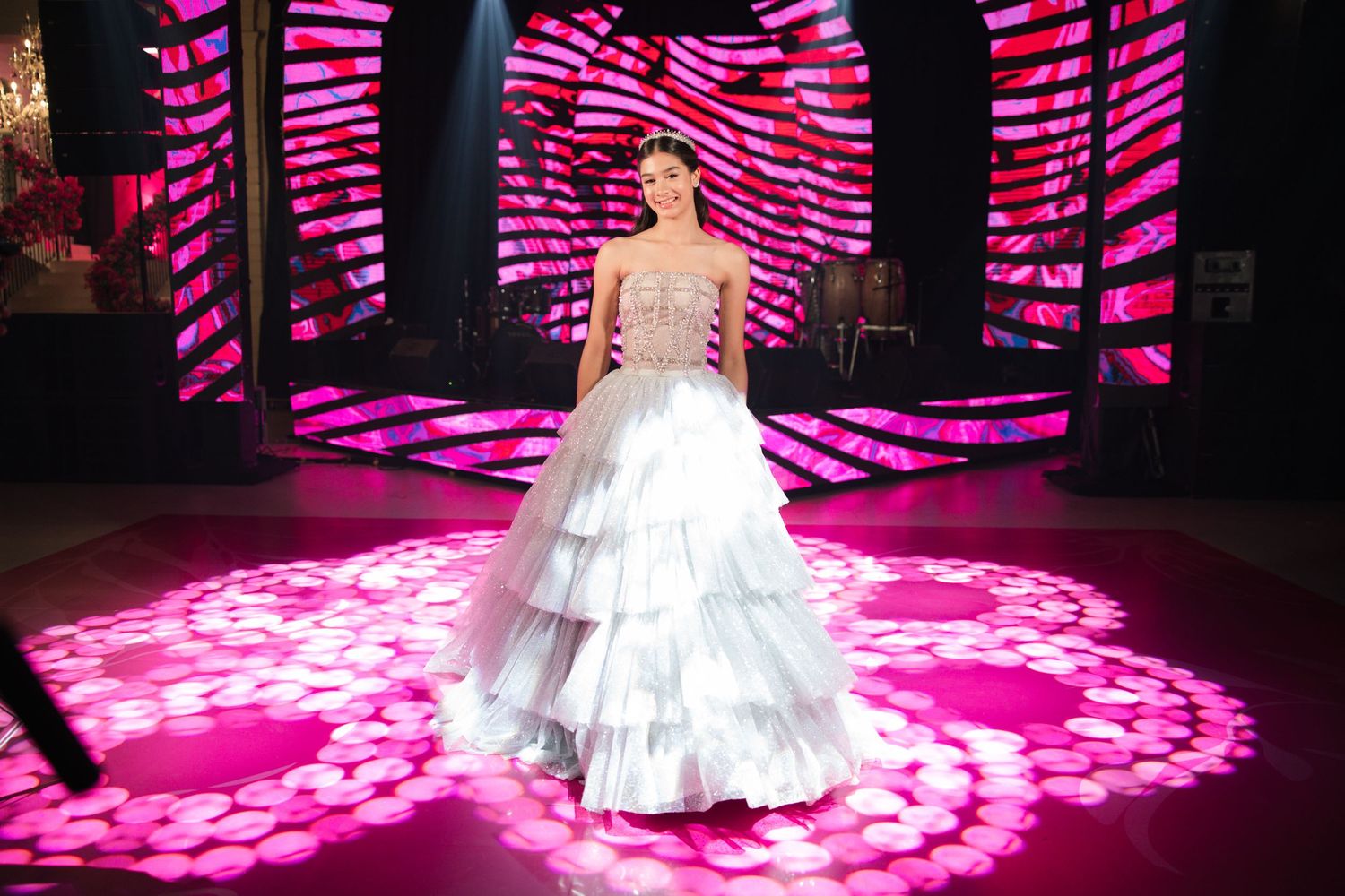 Person in tiered white ball gown stands on pink-lit stage with geometric light projections.