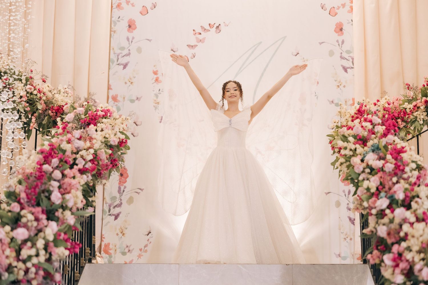 A bride in a white ballgown stands with outstretched arms surrounded by pink and white floral arrangements.