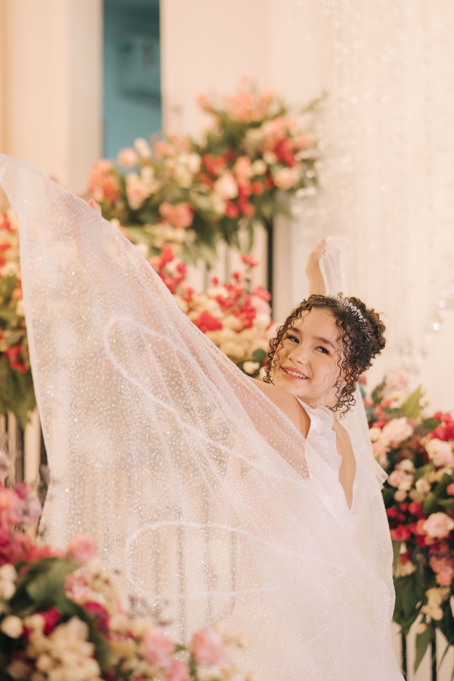 A bride holds her dress playfully while posing against a backdrop of coral and pink flowers.