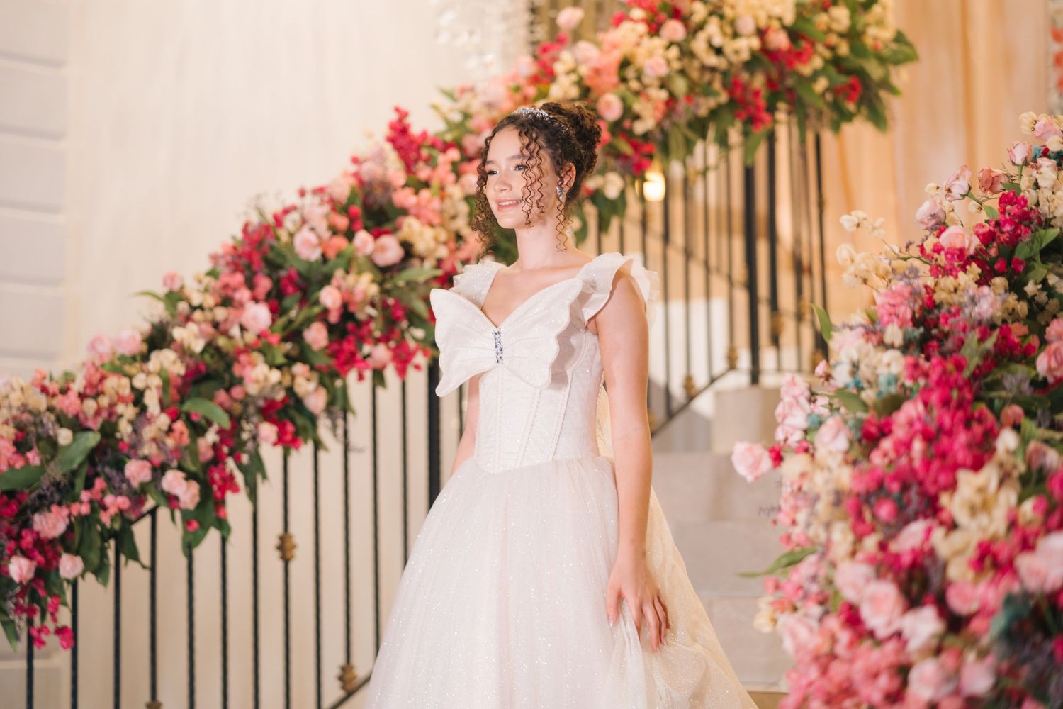 A bride in a white wedding gown stands by an elegant staircase adorned with pink and coral floral arrangements.