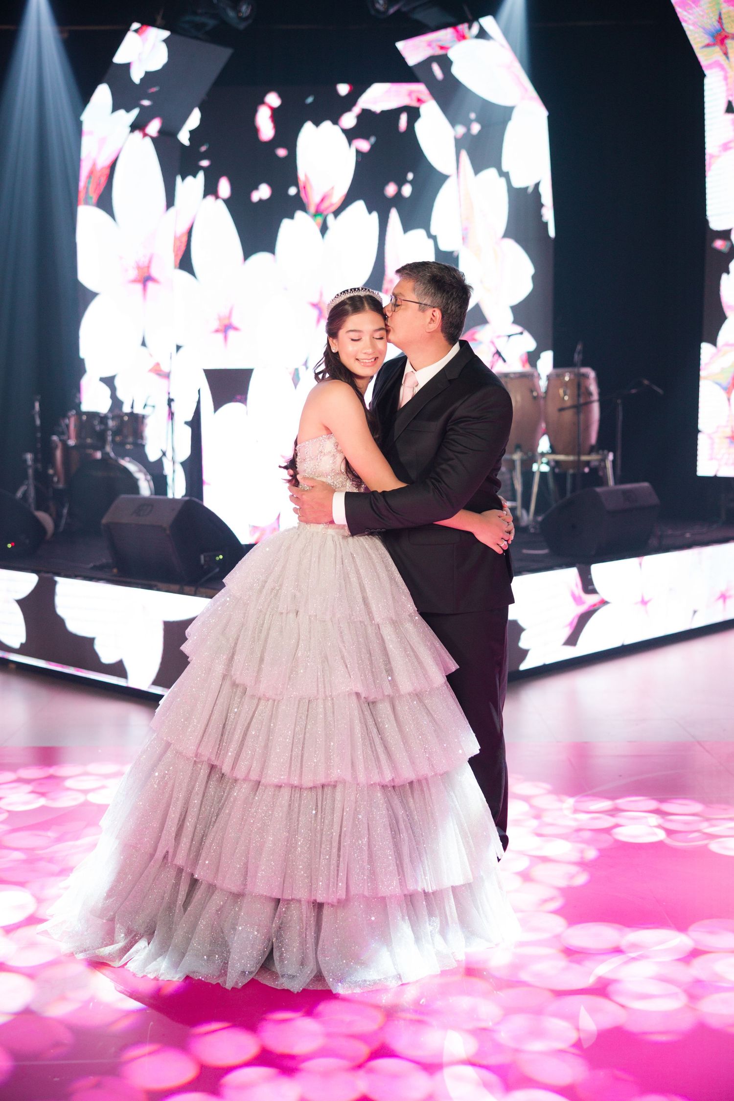 A couple shares a romantic moment on an illuminated pink dance floor under glowing white lanterns.