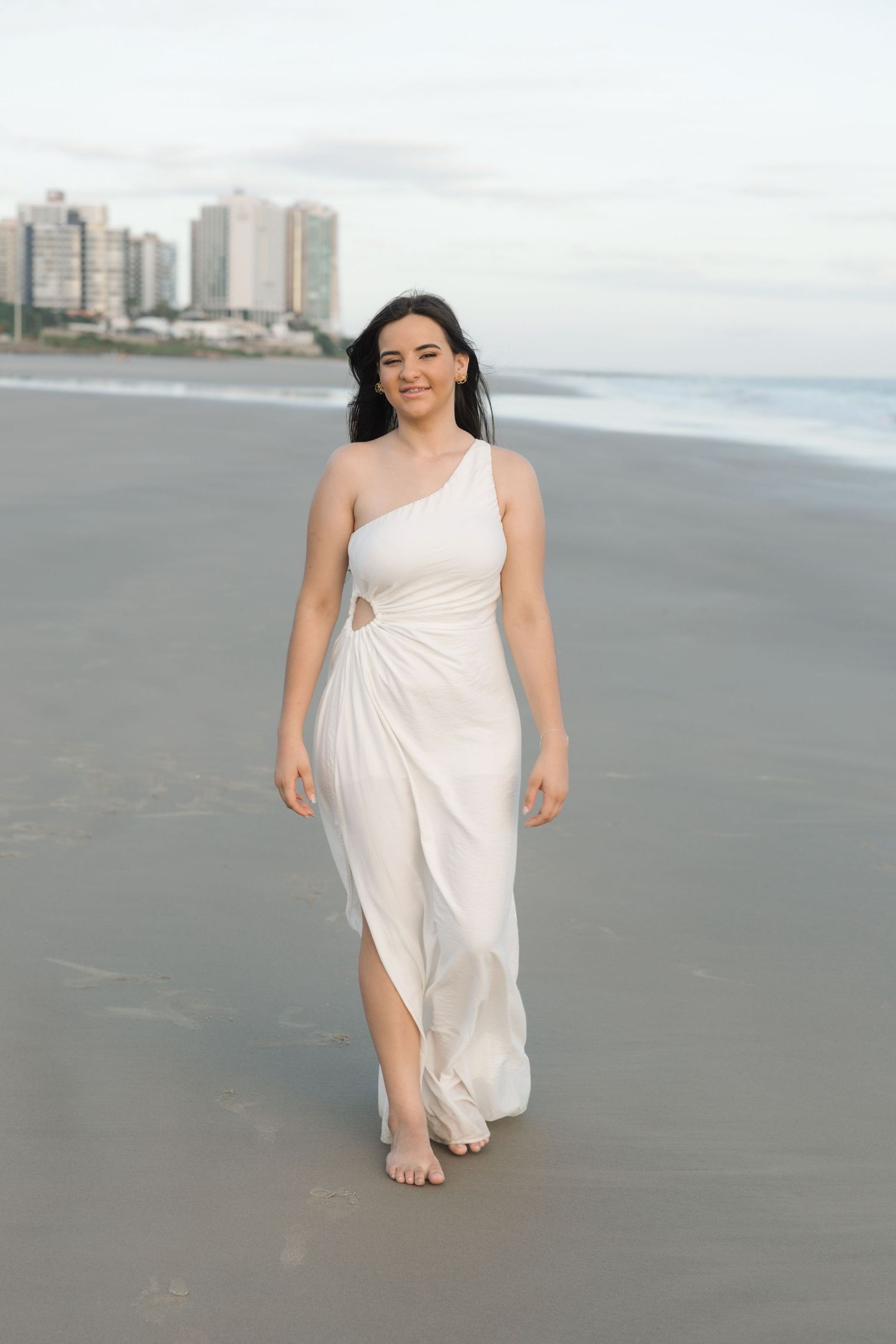 A person in a flowing white one-shoulder dress walks barefoot along a beach with city buildings visible in the background.