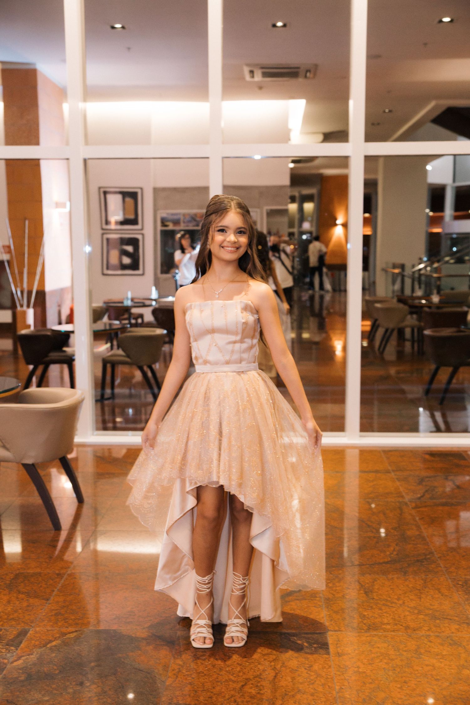 A formal high-low gown in rose gold sparkles photographed in a luxurious restaurant interior.