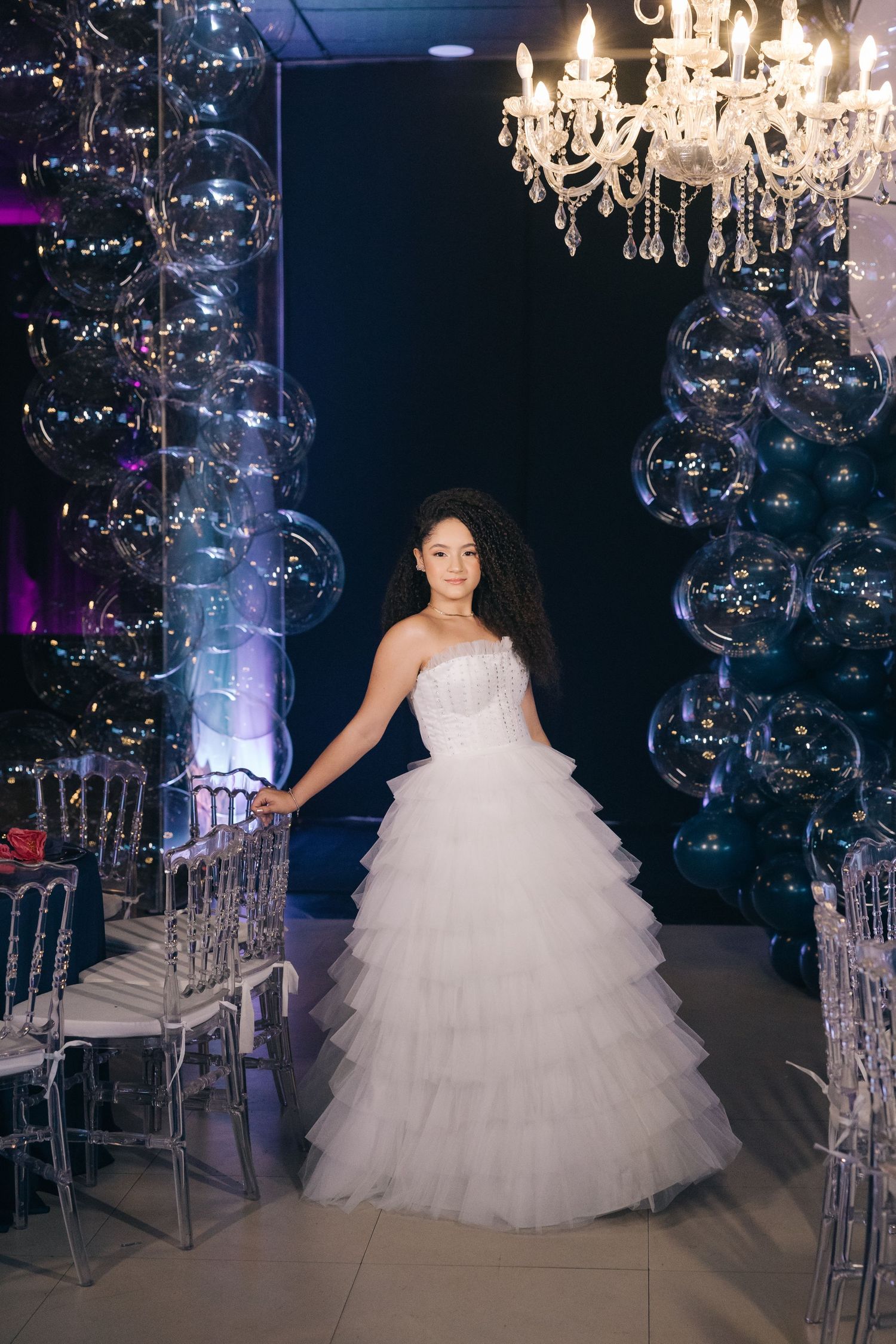A person in a layered tulle wedding gown stands between bubble decorations and clear chairs under a crystal chandelier.