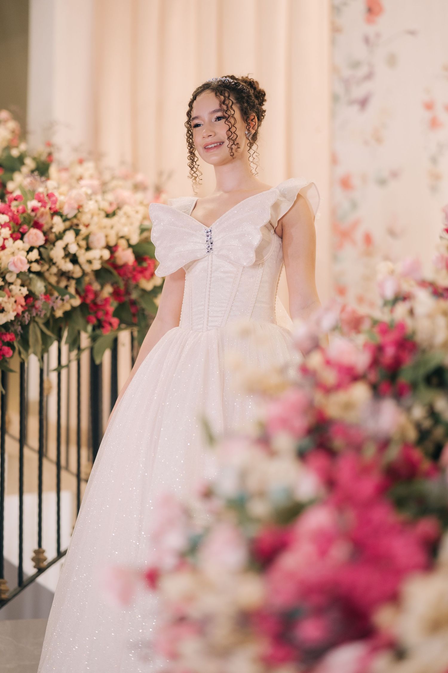 Romantic bridal portrait with white gown against pink floral arrangements and beige backdrop.