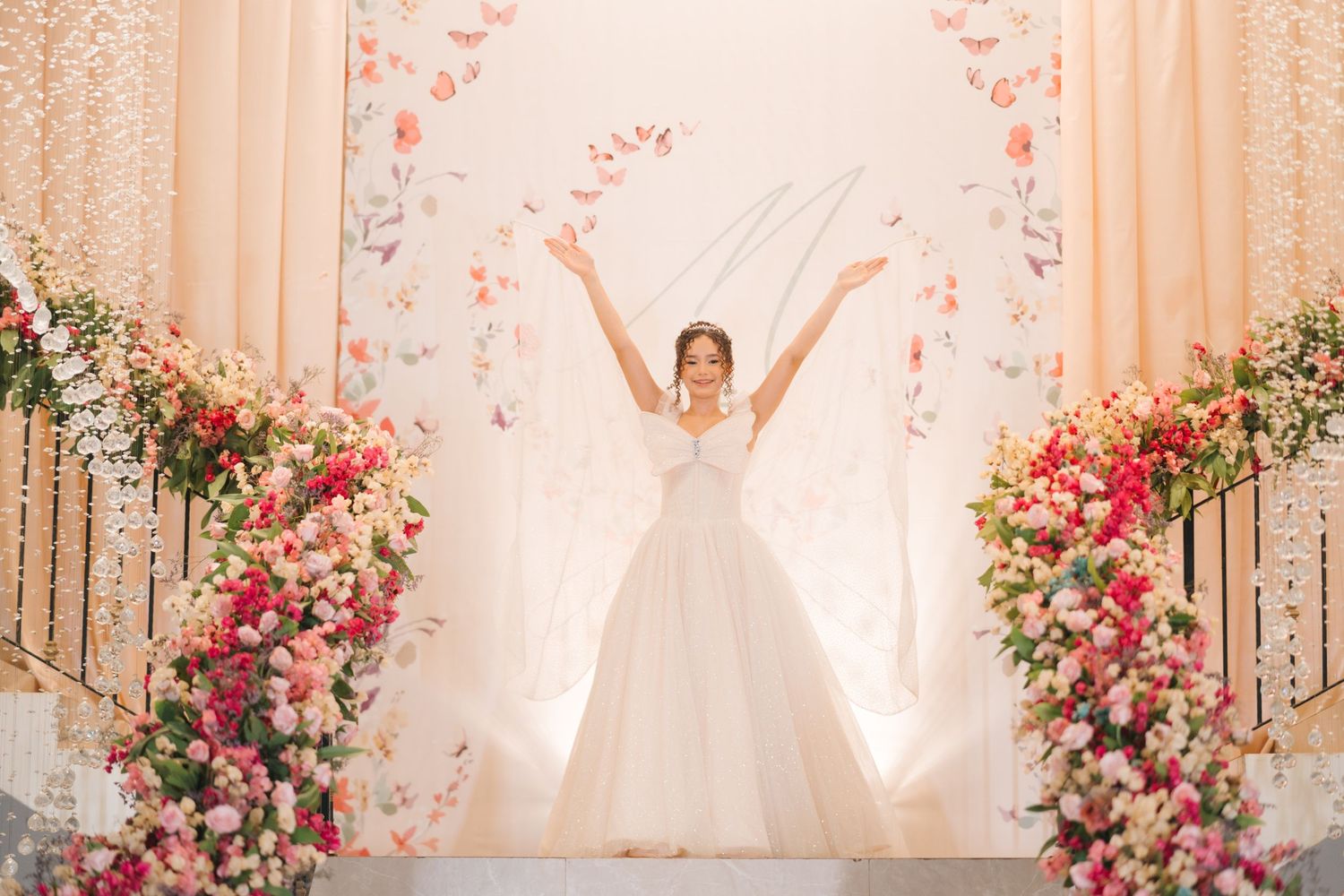A joyful bride in a white ballgown stands between pink floral arrangements against an ivory backdrop.