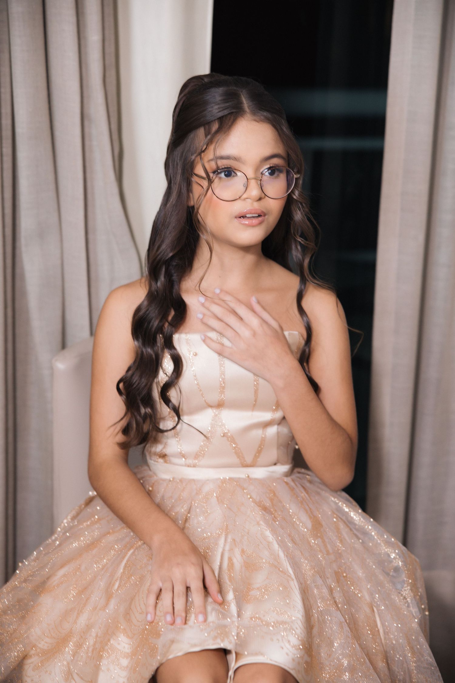 Young person in round glasses and flowing blush-colored gown poses elegantly against cream curtains.