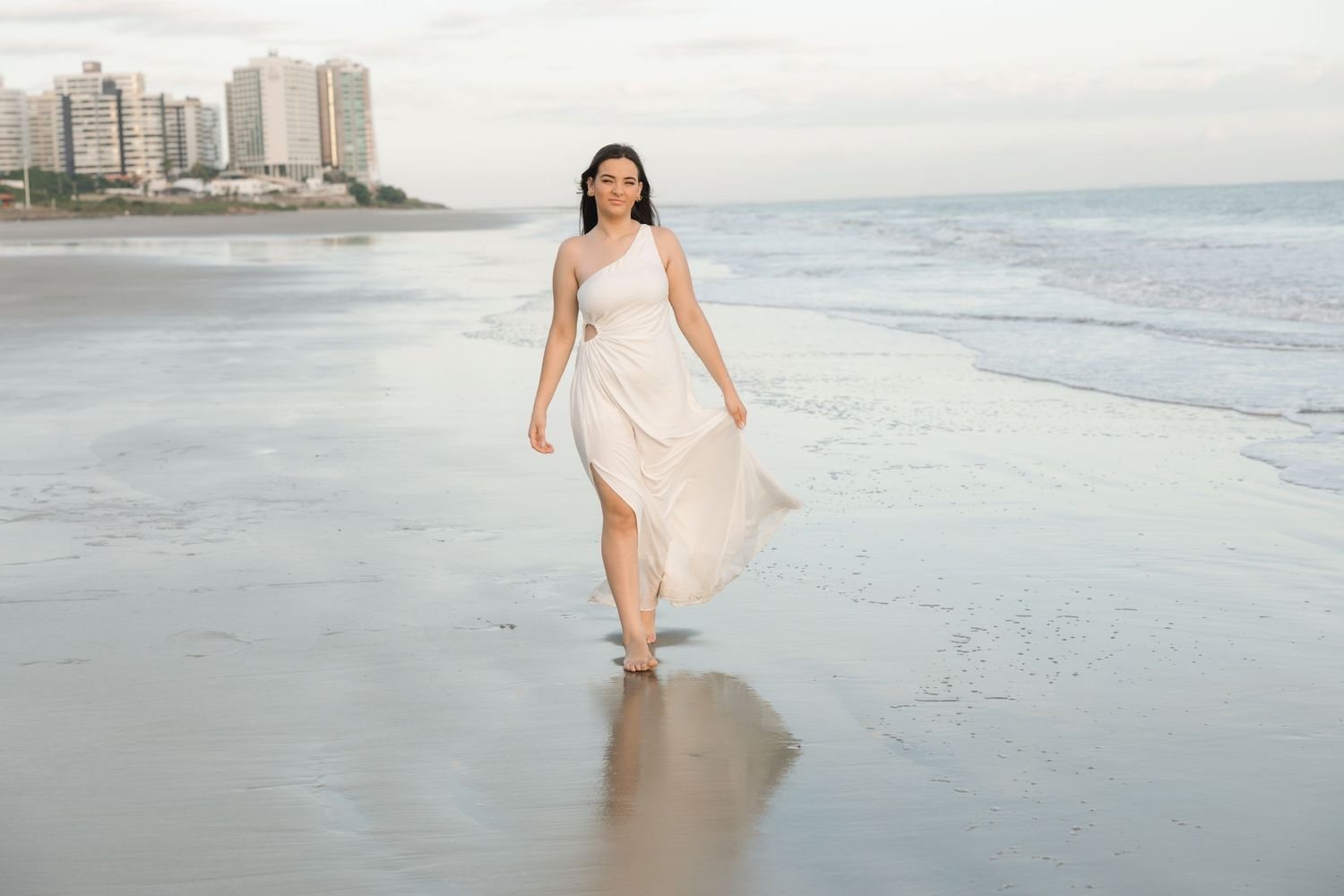 A person in a flowing white sundress walks barefoot along a misty beach with high-rise buildings in the background.