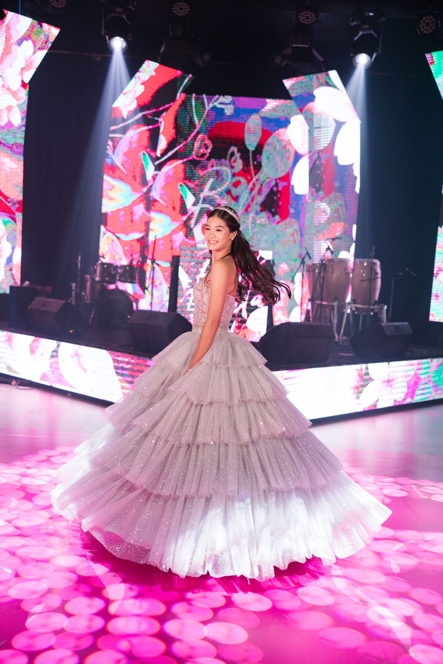 Person twirls in a layered tulle gown on illuminated pink floor with colorful digital backdrop.