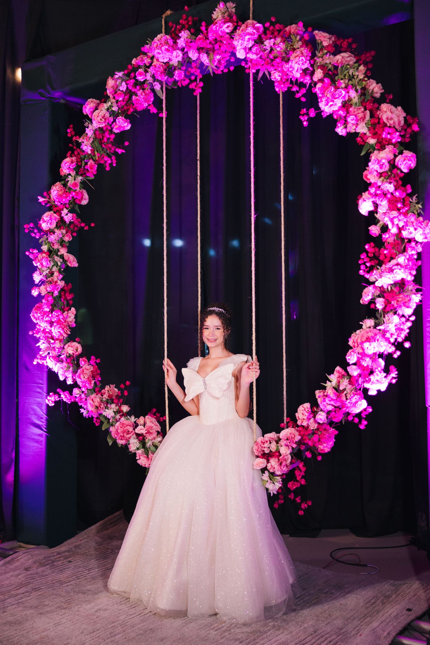 A bride poses on a swing surrounded by a pink floral arch with dramatic lighting.