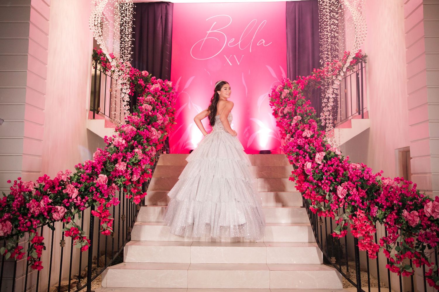 Grand staircase decorated with pink roses and lights features a person in a white ball gown.