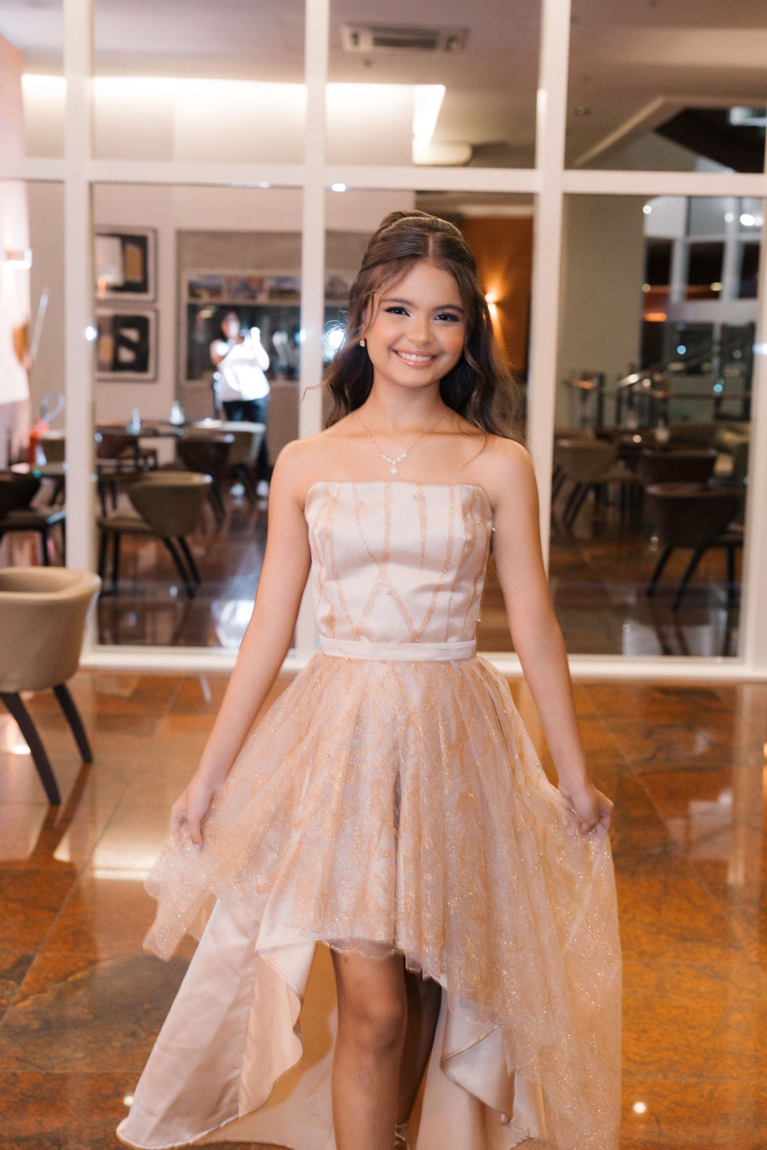 A person in a strapless high-low blush gown stands elegantly in a restaurant interior with warm lighting and glass walls.