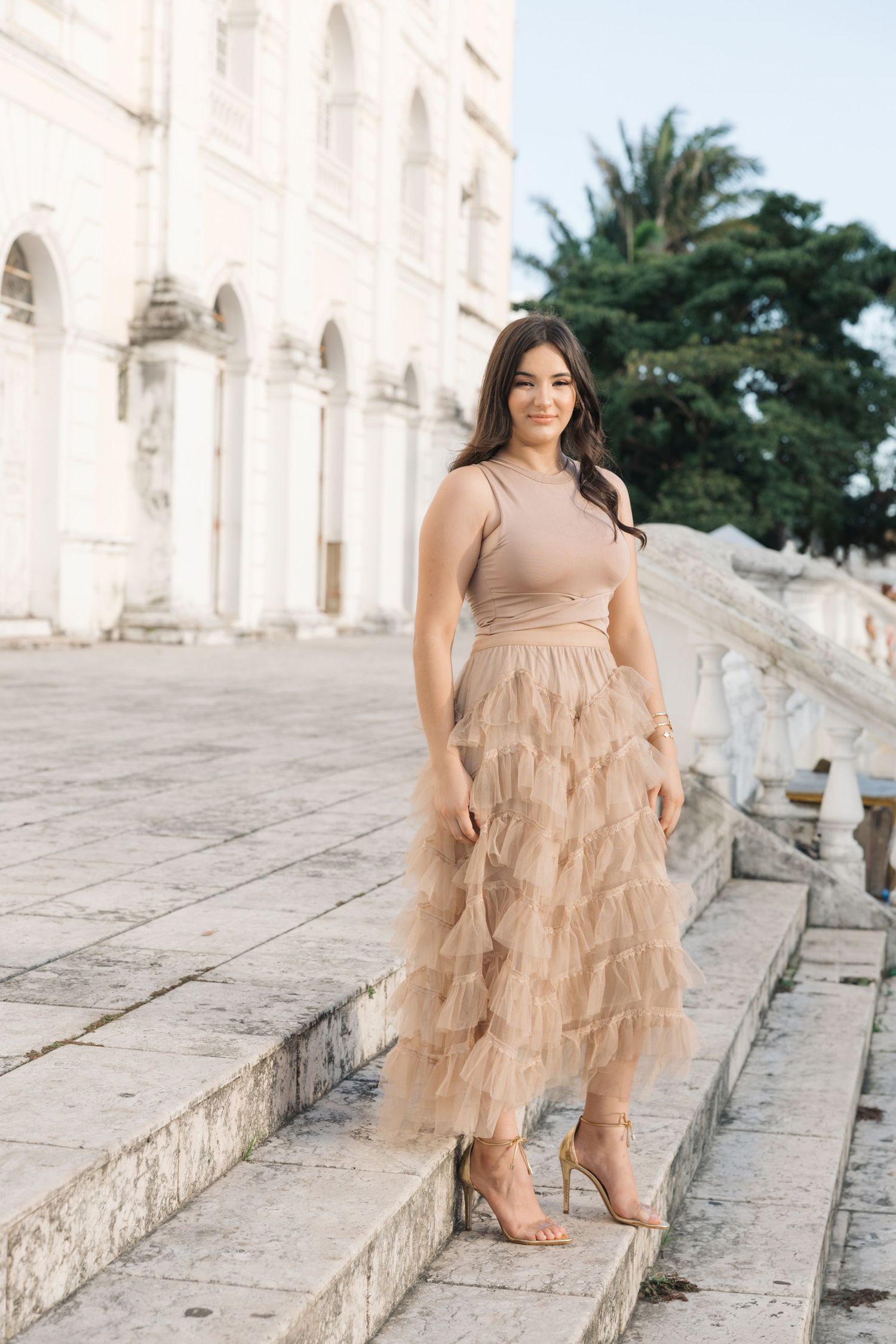 Elegant ruffled champagne tulle dress and heels worn on steps of historic white stone building with palm trees.