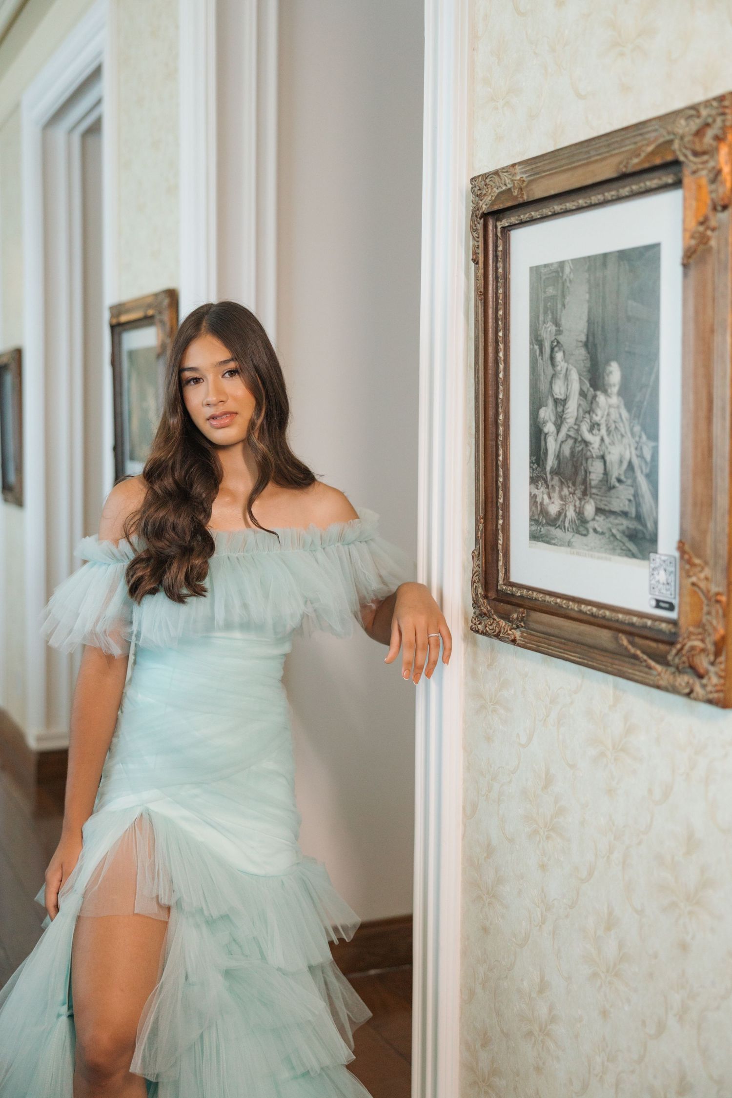 An off-shoulder mint green dress with delicate tulle details is styled in a classic interior hallway with framed artwork.