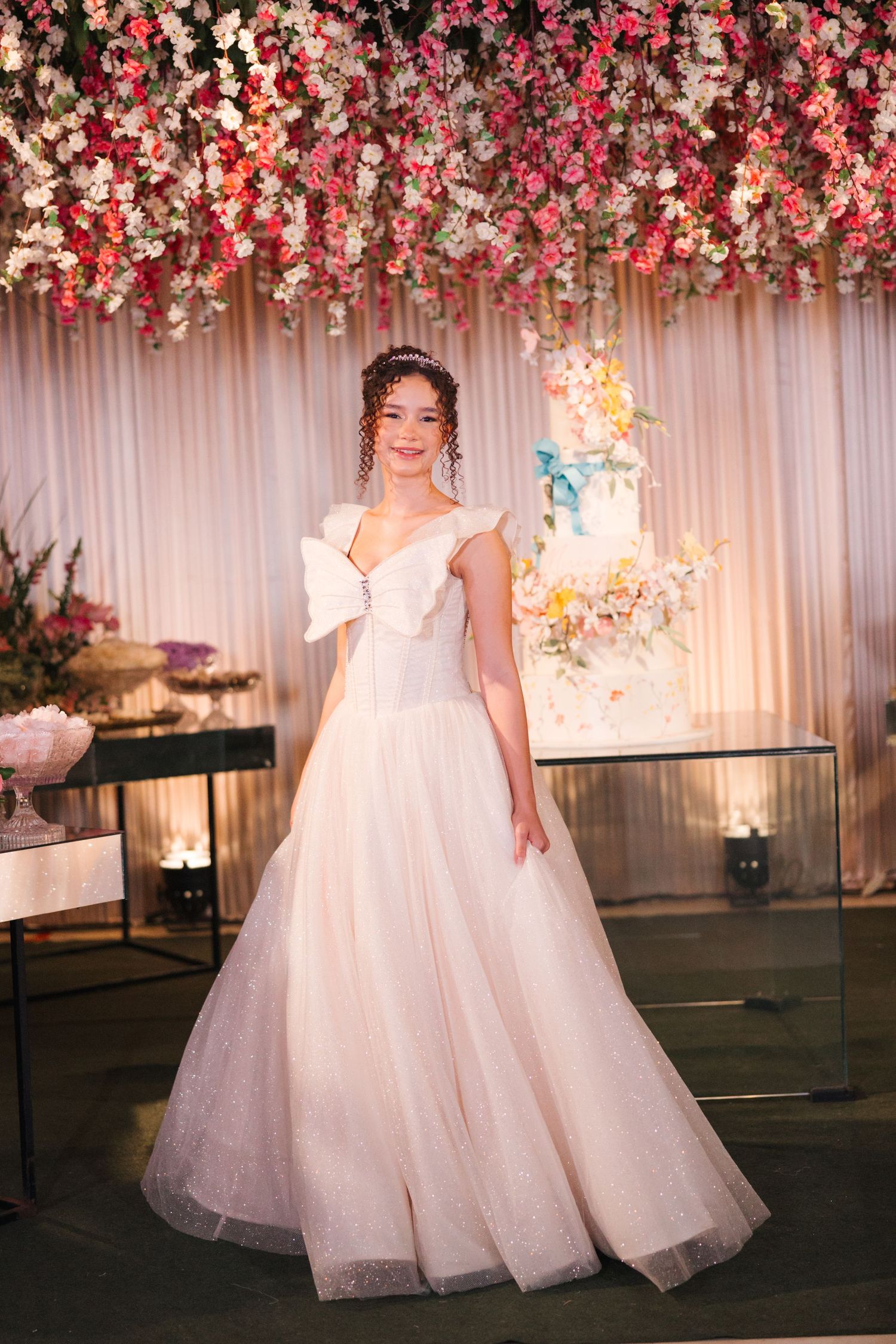 A bride wearing an off-shoulder wedding gown poses in front of a romantic floral wall and cake display.