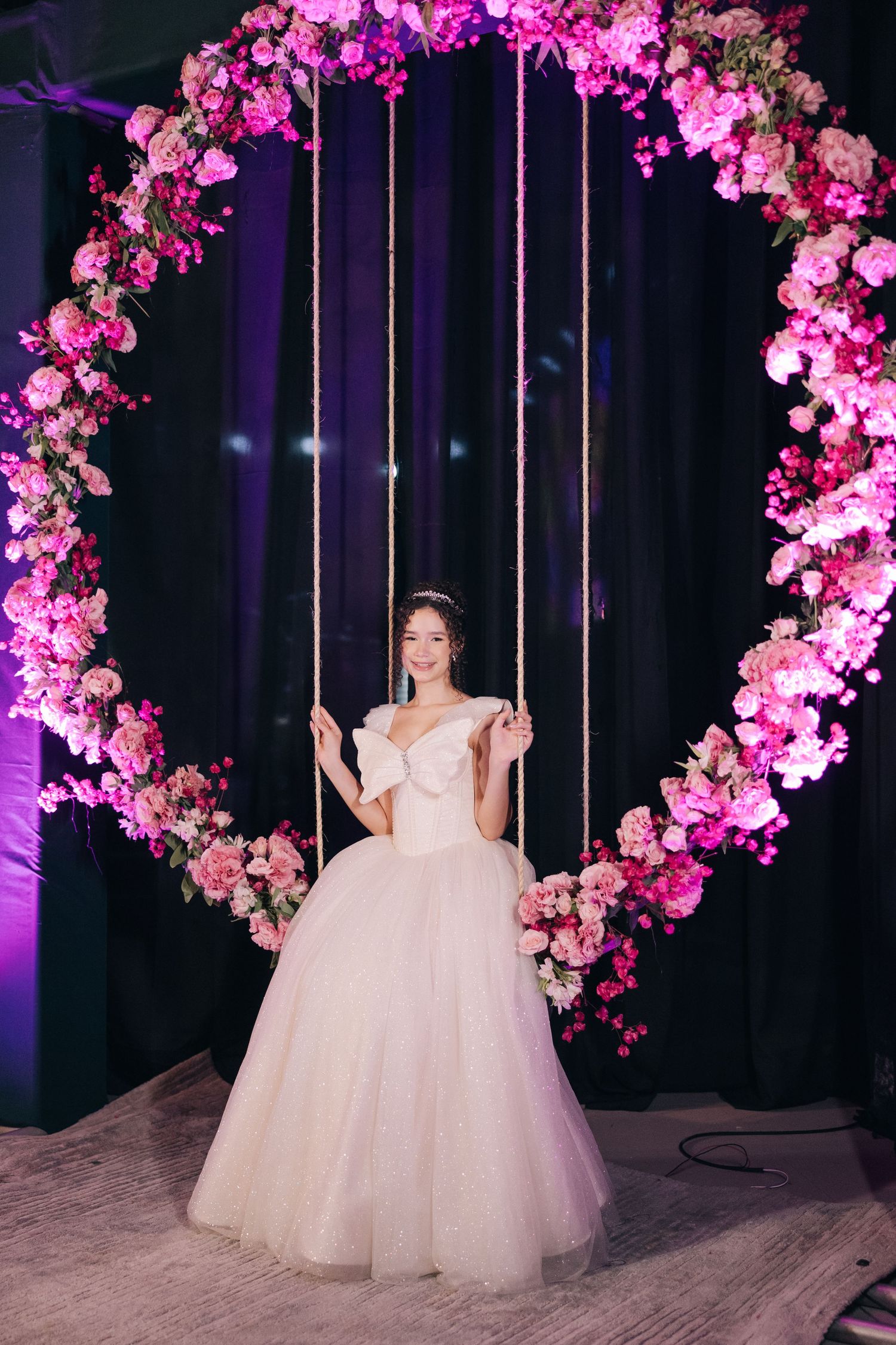 Elegant ballgown-clad figure on a suspended swing decorated with a pink floral arch in a dramatic purple-lit setting.