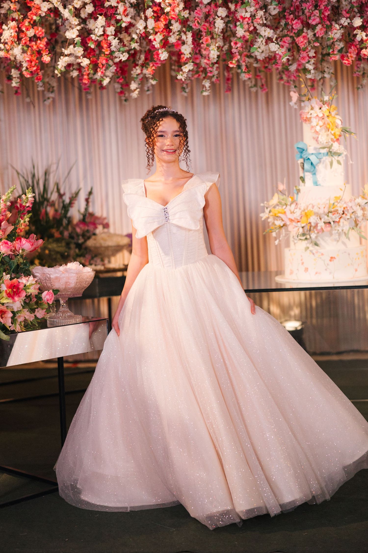 A bride in a tulle ballgown stands before a floral backdrop with pink and coral flowers.