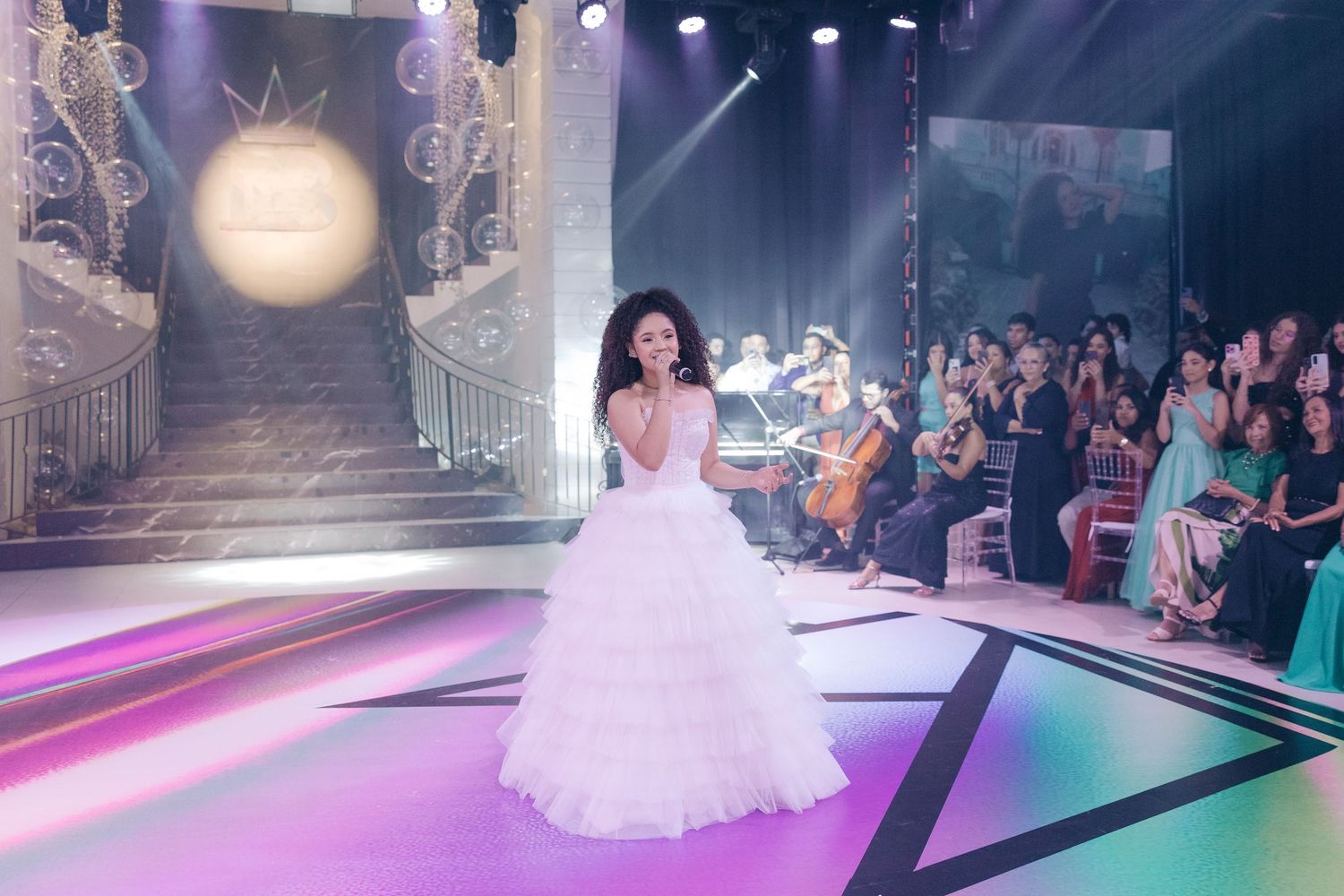 Person in a flowing white ball gown performs on an illuminated runway stage with audience members seated in background.