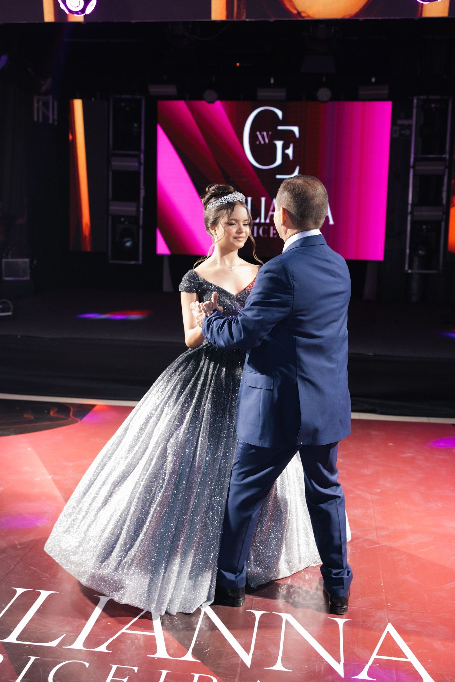 A couple dances on stage in formal attire with a glittering ball gown and navy suit.