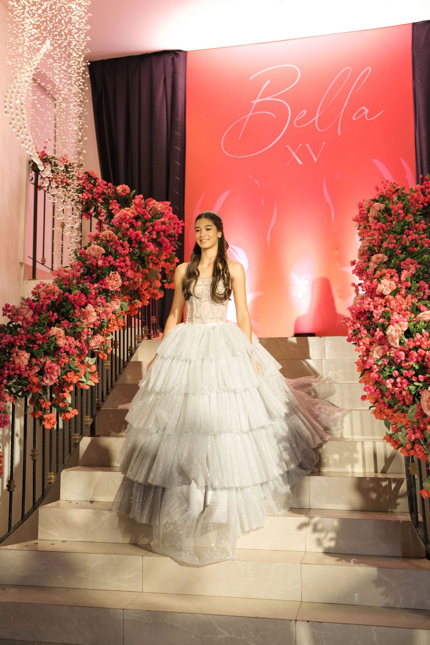 Elegant tiered ballgown photographed on stairs with pink floral decorations and 'Bella XV' signage.