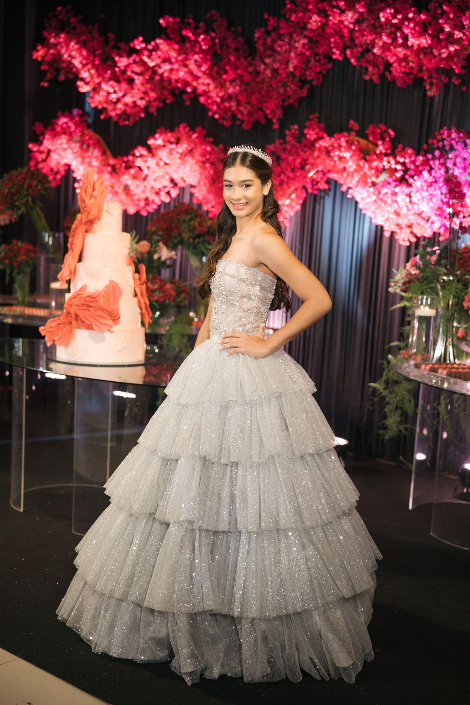 A shimmering silver tiered ball gown with sparkles stands before a backdrop of pink cherry blossoms and a wedding cake.